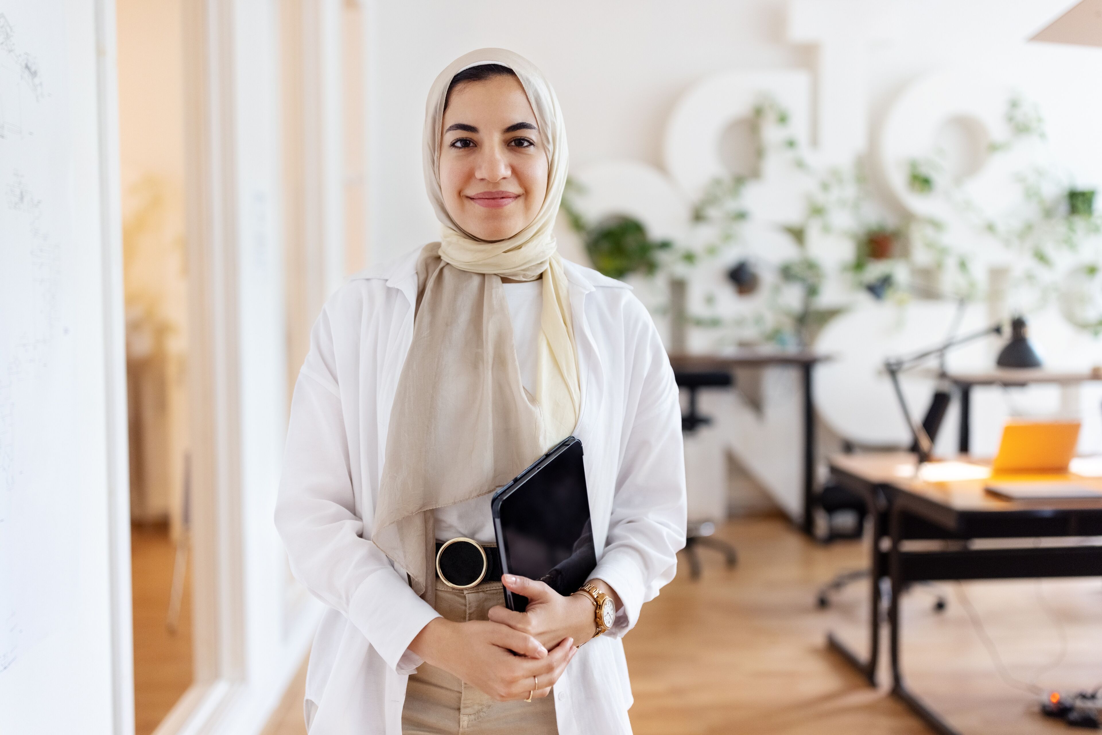 Portrait of happy young Muslim businesswoman holding digital tablet. Female entrepreneur with a headscarf in office looking at camera and smiling.
