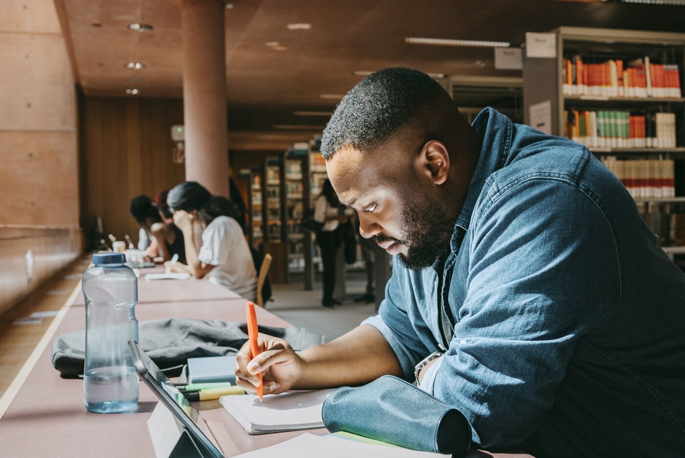 Student writing in notebook while sitting with tablet PC in college library