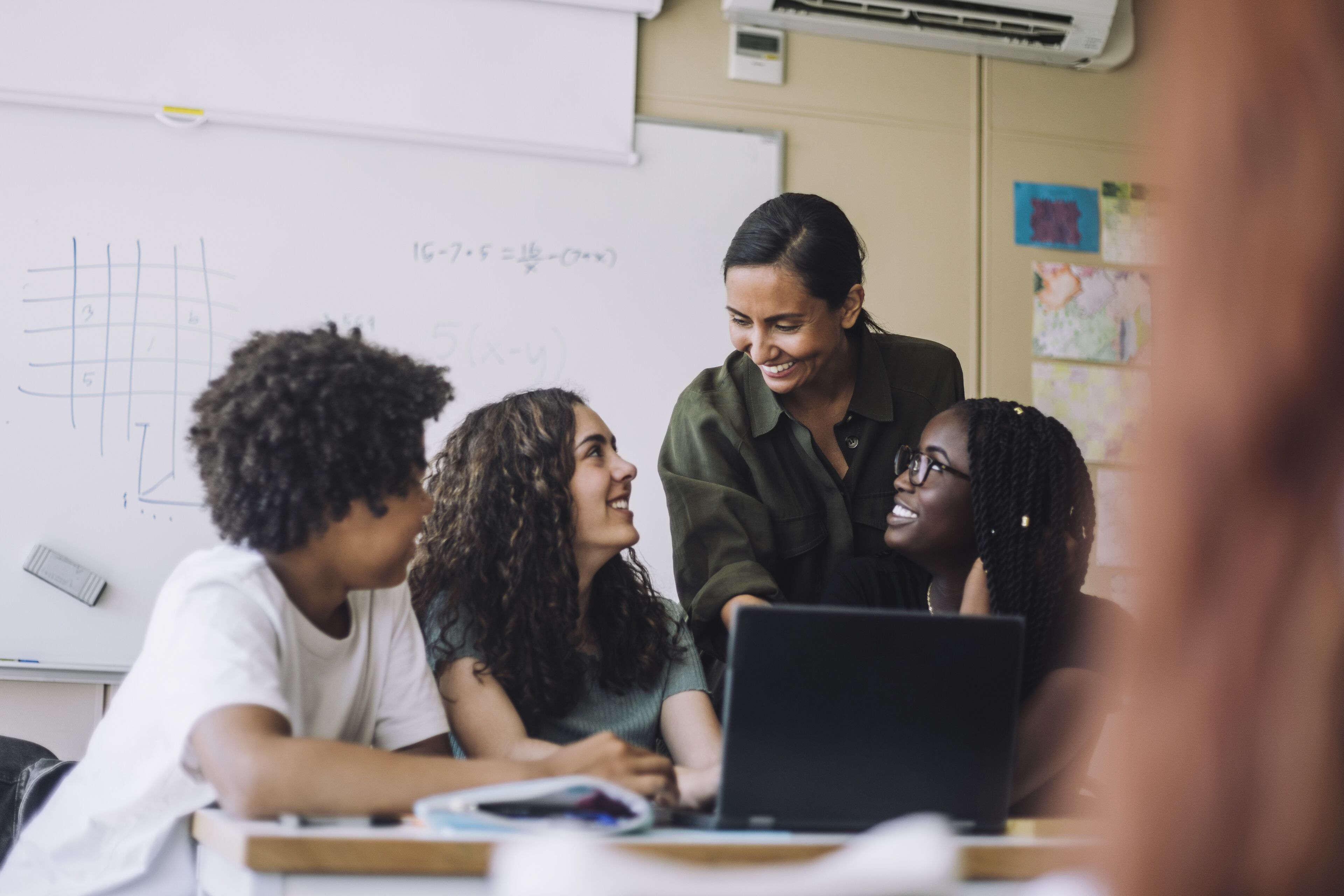 A teacher engaging with three students around a laptop in a classroom, with a math problem on the whiteboard in the background.