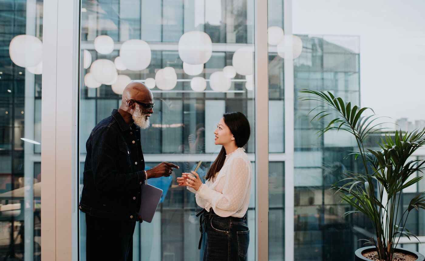 An older man and a young woman stand in an office building beside a huge window and converse.