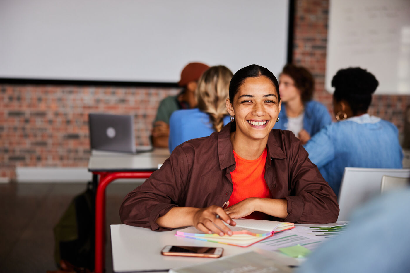 A smiling young woman sits at a table in a classroom, surrounded by other students, holding a notebook and looking directly at the camera.