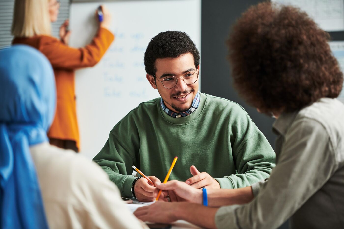 A young man in a green sweater sits at a table, engaging in conversation with two other individuals while a woman in an orange sweater stands in the background writing on a whiteboard.