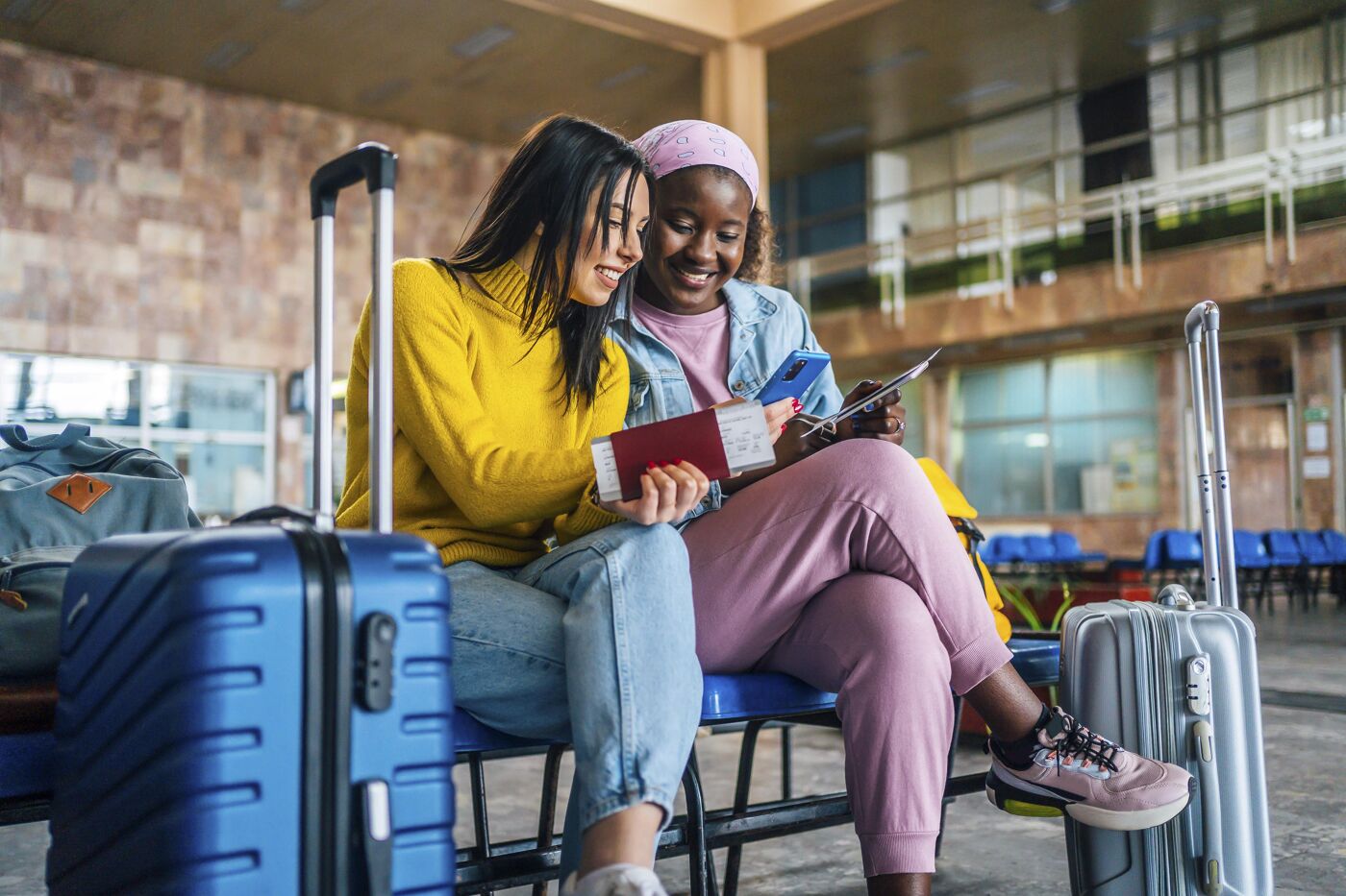 Two women sitting in an airport lounge review their travel documents and smile. Surrounded by luggage, they appear excited about their journey. One wears a yellow sweater, the other a denim jacket with a pink headscarf.