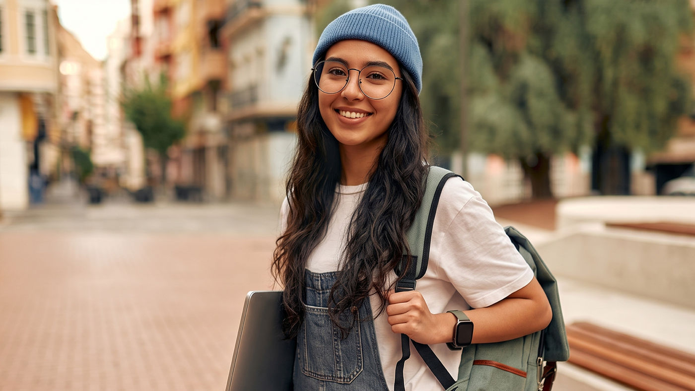 A young woman wearing a blue beanie and glasses smiles at the camera, carrying a green backpack and a laptop. She is outdoors, possibly near an urban street.