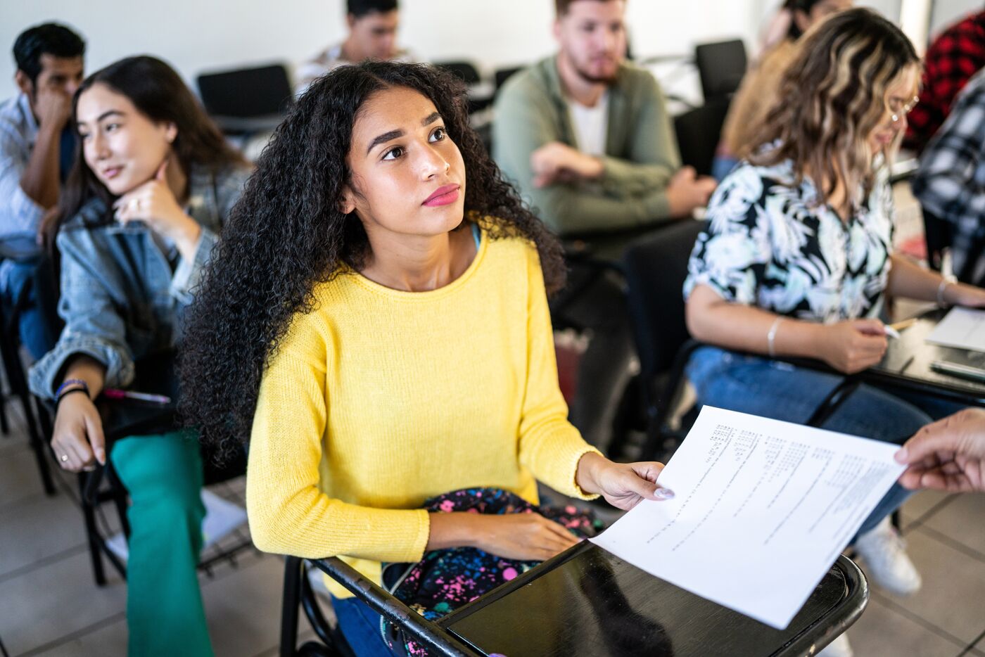 A focused student in a yellow sweater examines a paper during a class discussion, surrounded by fellow learners.