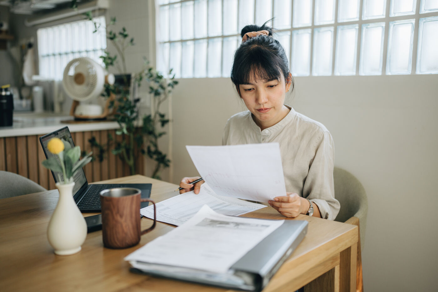 A woman sits at a wooden table, reviewing paperwork while a laptop and a mug sit beside her.