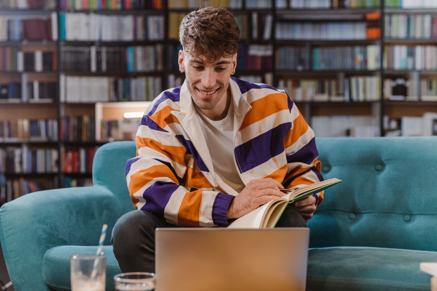 A young man sits on a teal couch in a library, holding an open book and looking at a laptop.