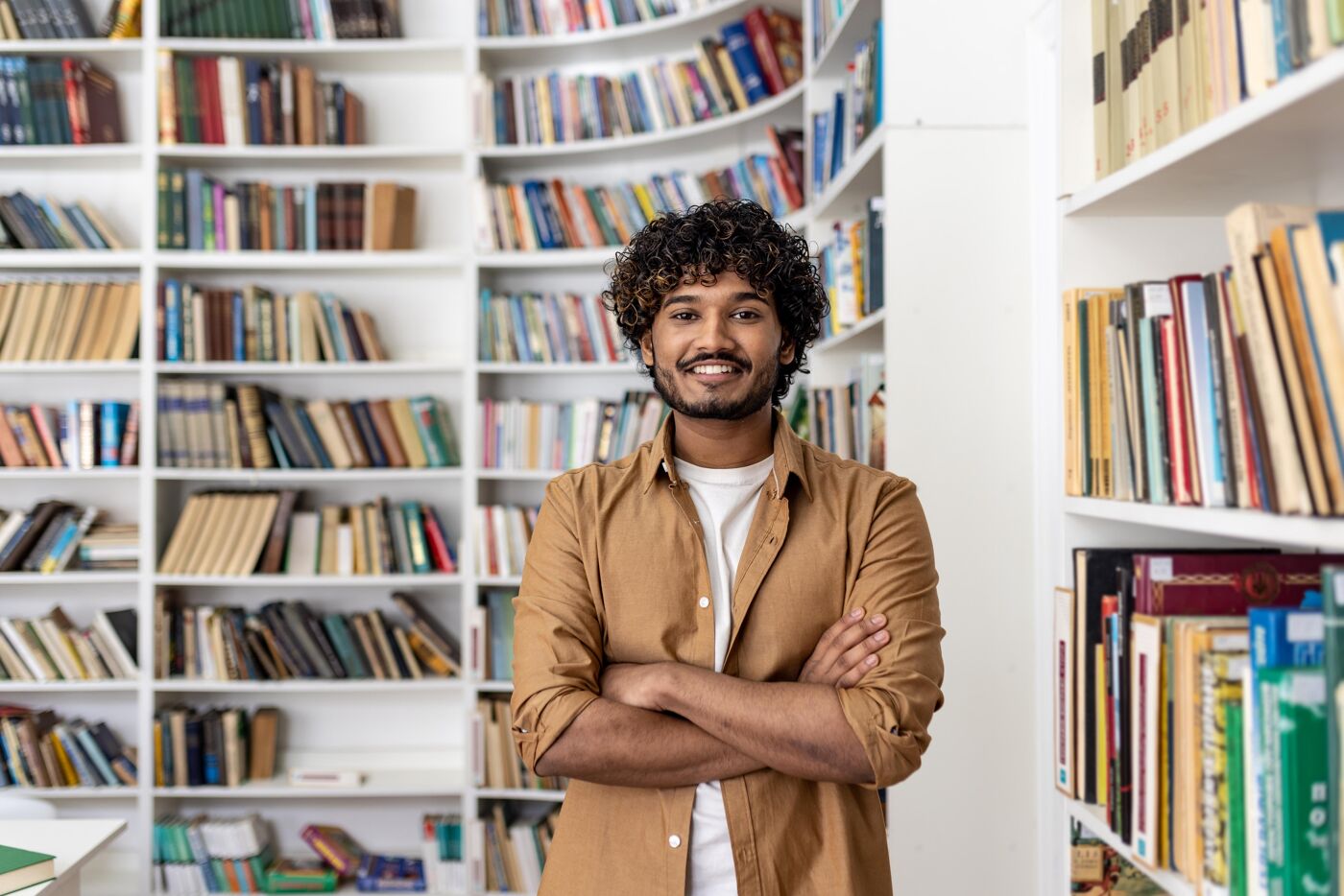 A smiling young man with curly brown hair stands with his arms crossed in front of bookshelves filled with various books.