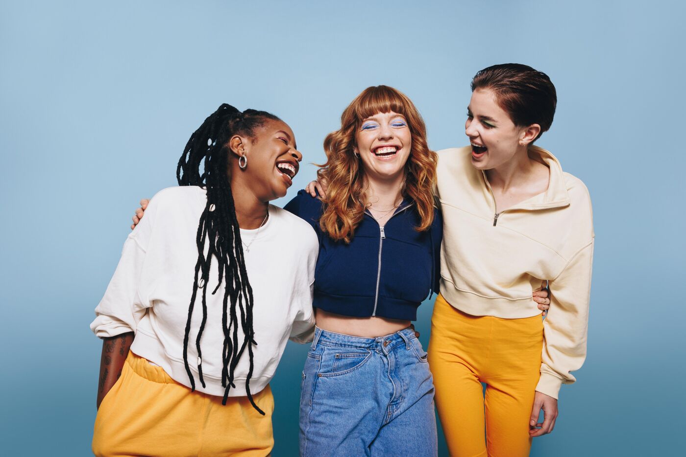 Three young women are standing close together, laughing and embracing. The woman on the left has dark skin and long braids, the woman in the center has red hair, and the woman on the right has fair skin and short hair. They appear happy and carefree.