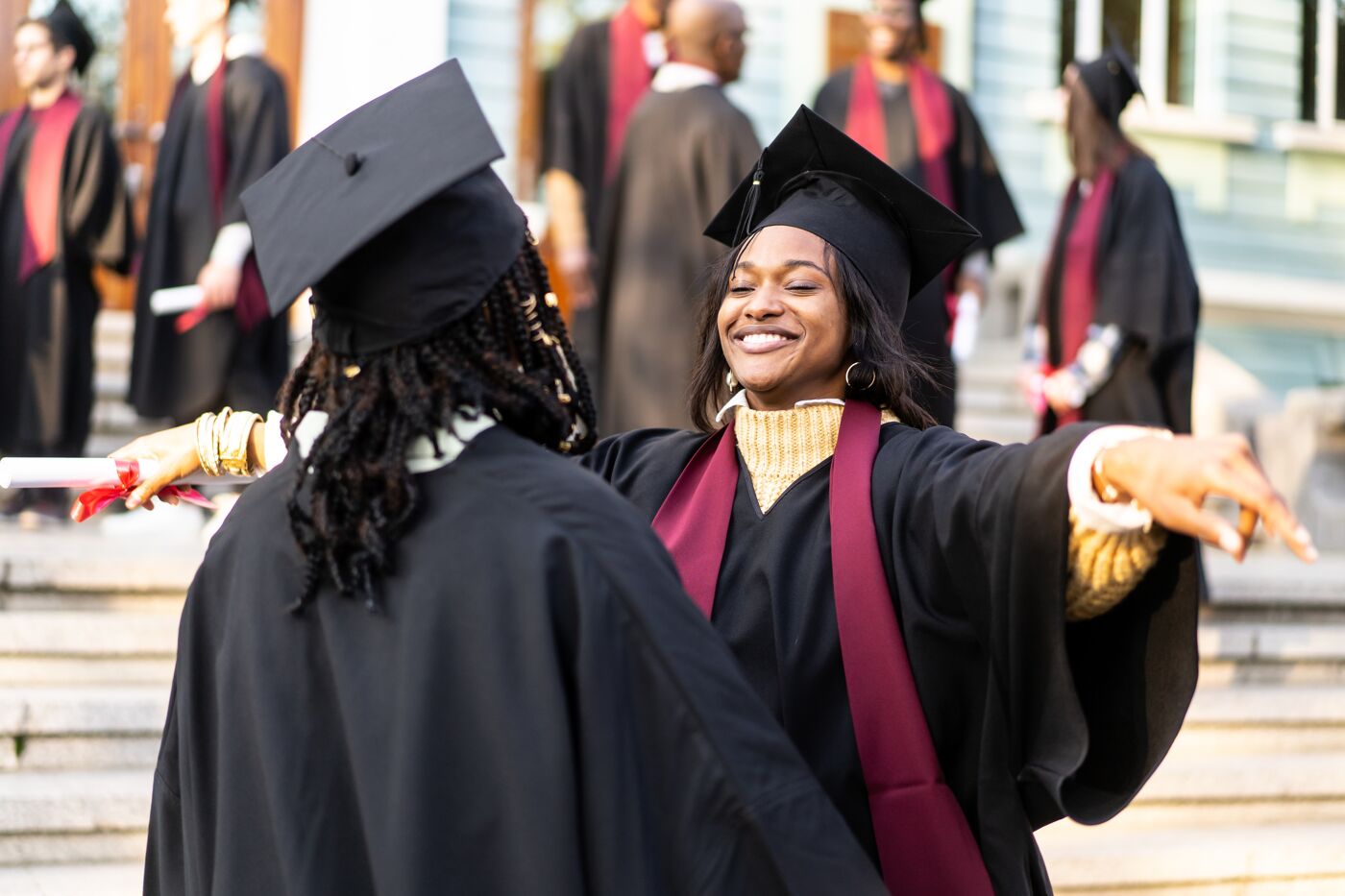 Two smiling women wearing graduation cap and gown looking at each other before they embrace on the university steps on graduation day