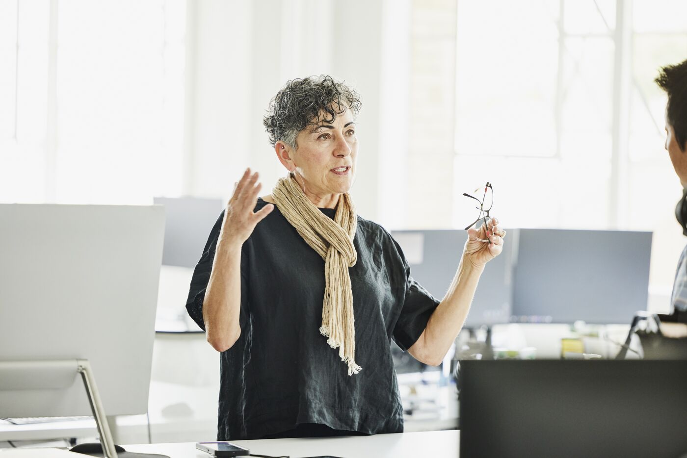 A professional woman conducts a presentation in an office, holding glasses and gesturing for emphasis.