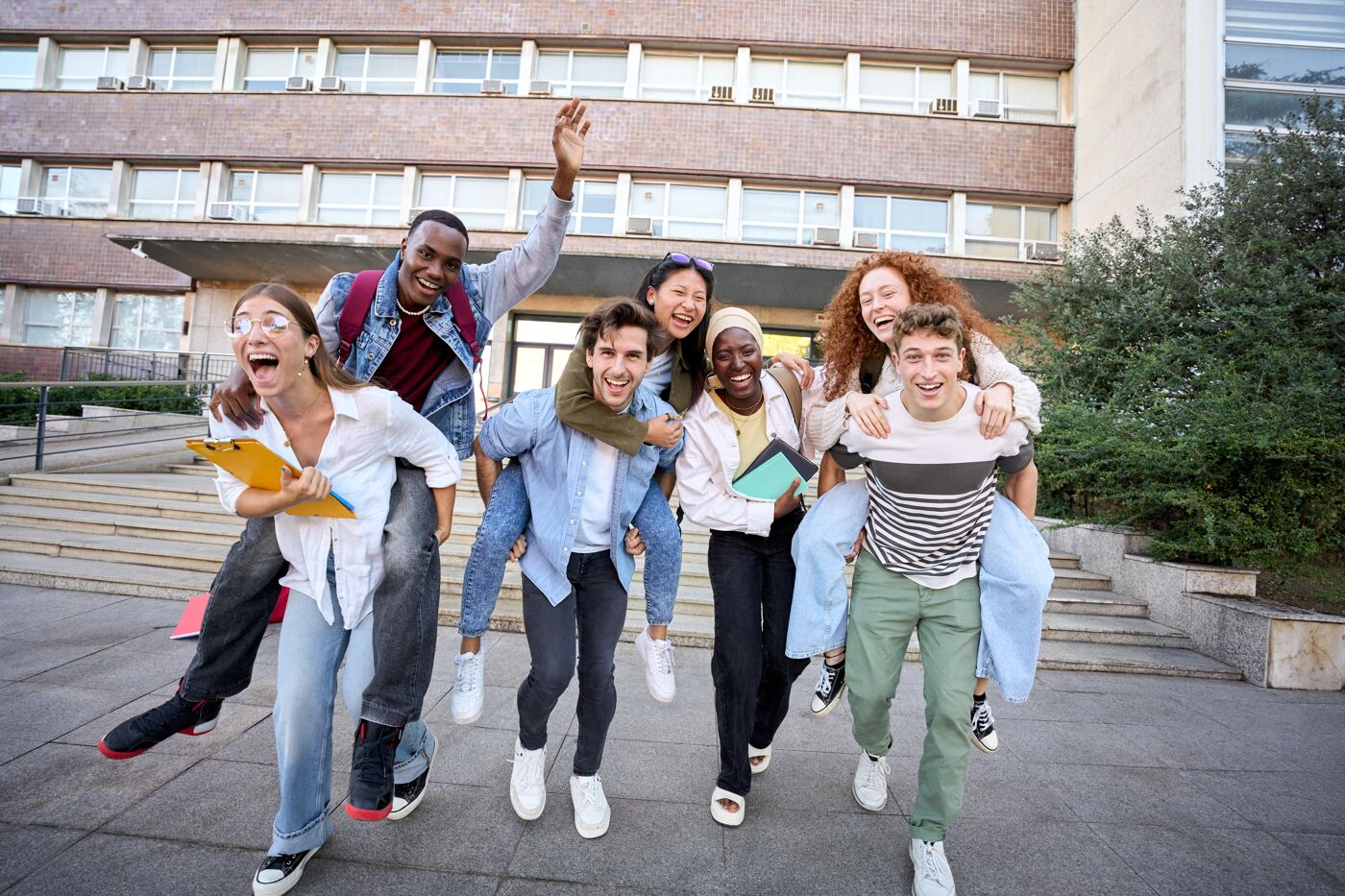 Multiracial group of student friends doing piggyback all together after school, having fun, looking at camera cheerfully.