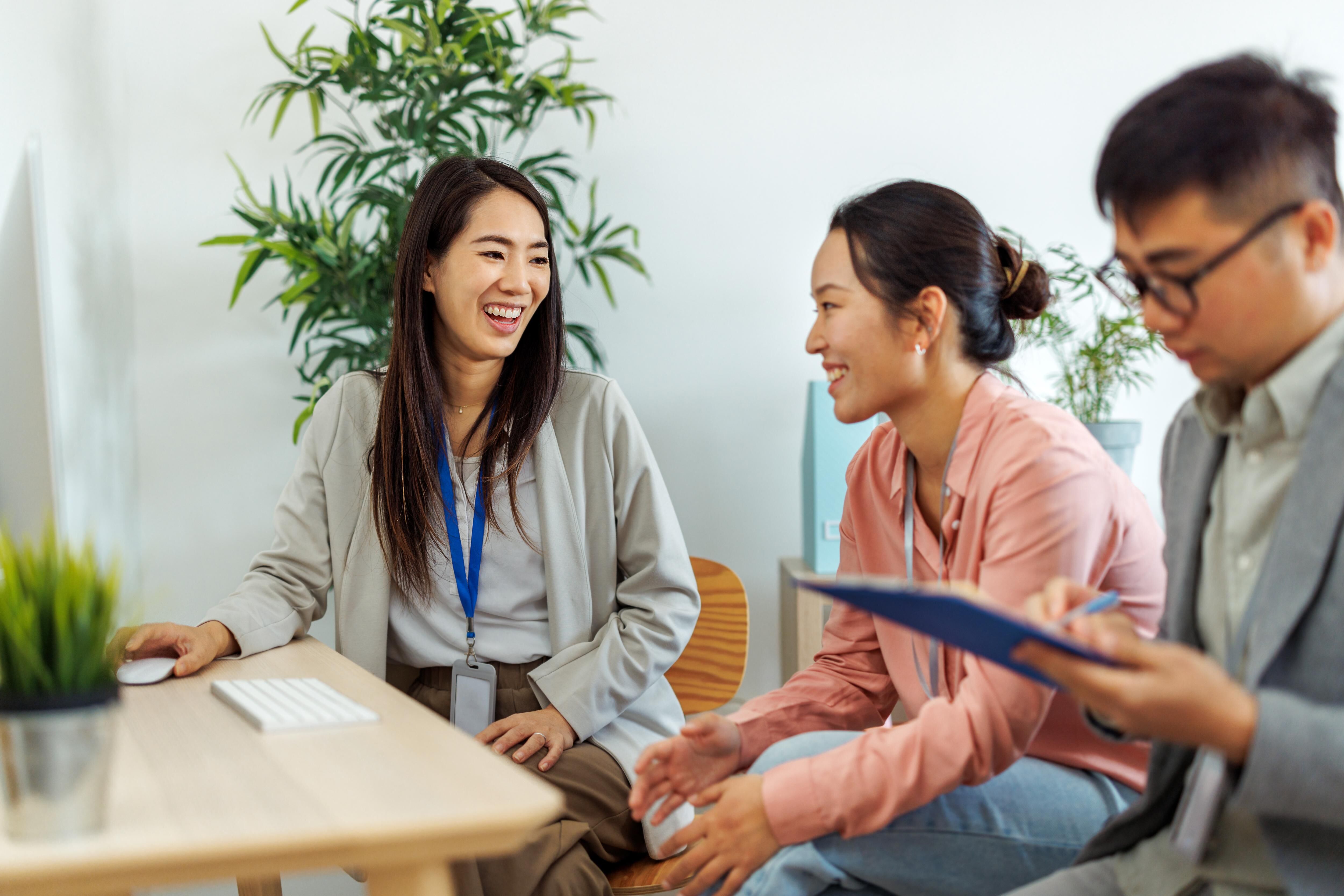 The image shows three people in an office setting. Two women are smiling and appear to be engaged in a conversation, while a man is looking down and holding a clipboard. There's a plant in the background.