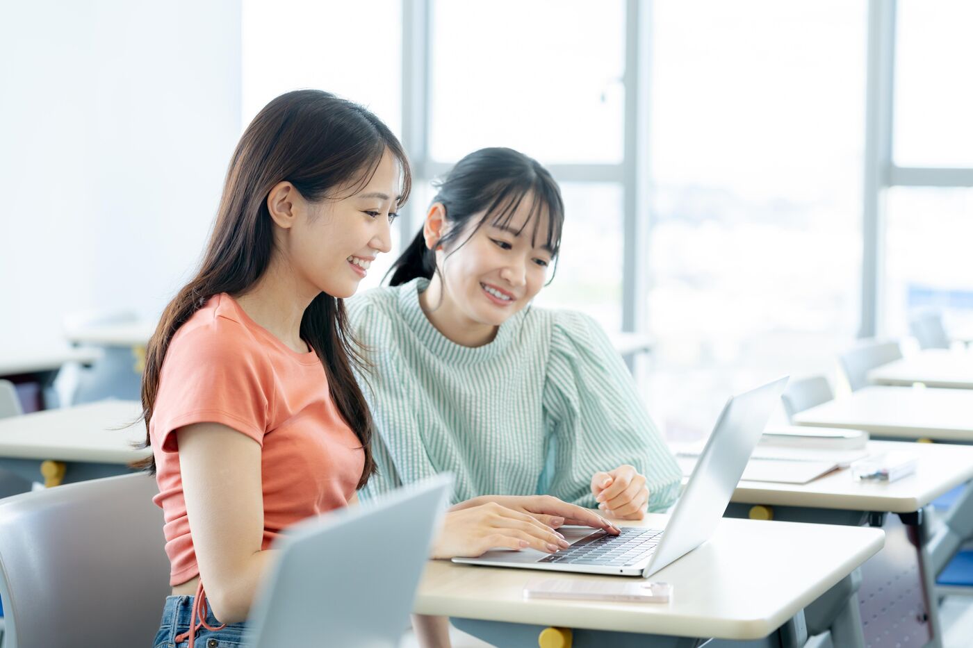 Two young women are sitting at a desk, looking at a laptop. They appear to be collaborating or working together on a project, with smiles suggesting a positive and engaging atmosphere.