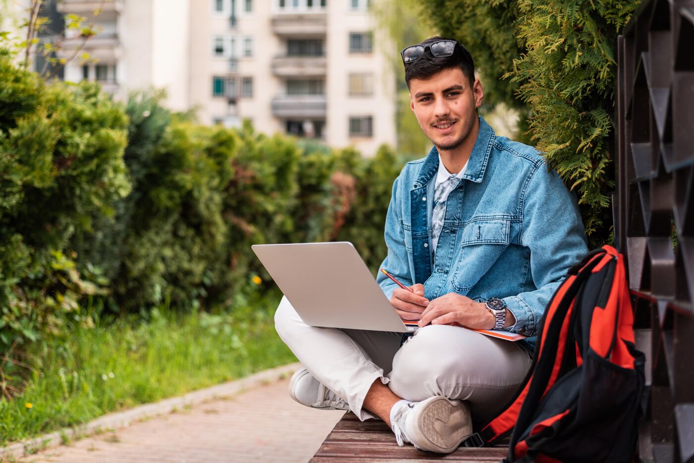A smiling young man with sunglasses on his head, sitting outdoors with a laptop and notebook, preparing to study.