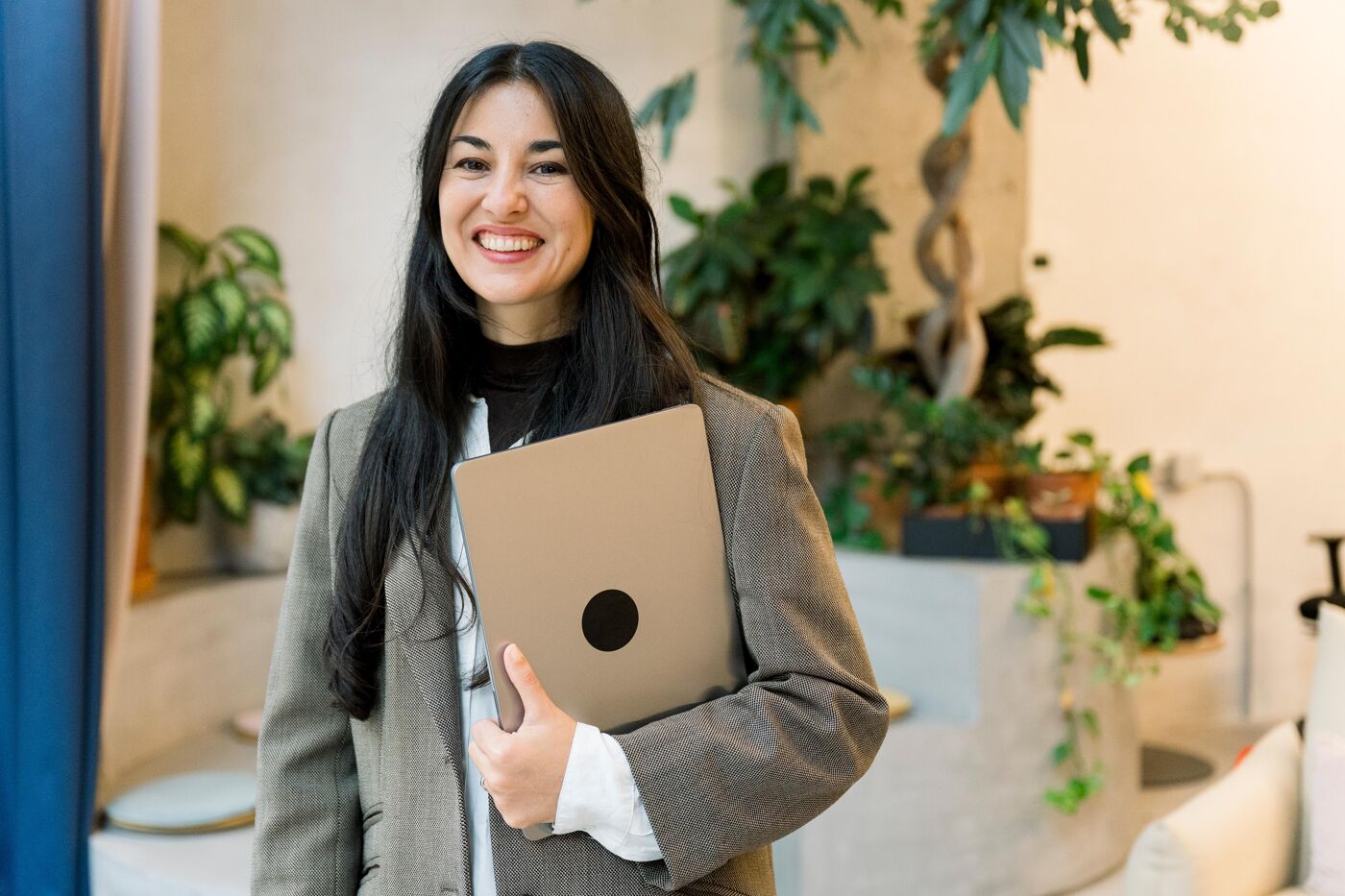 A smiling woman in a blazer and white turtleneck holds a laptop under her left arm, with green plants in the background.