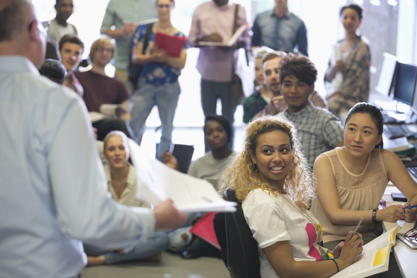 A vibrant classroom scene with a diverse group of students attentively listening to a lecturer who is holding papers.