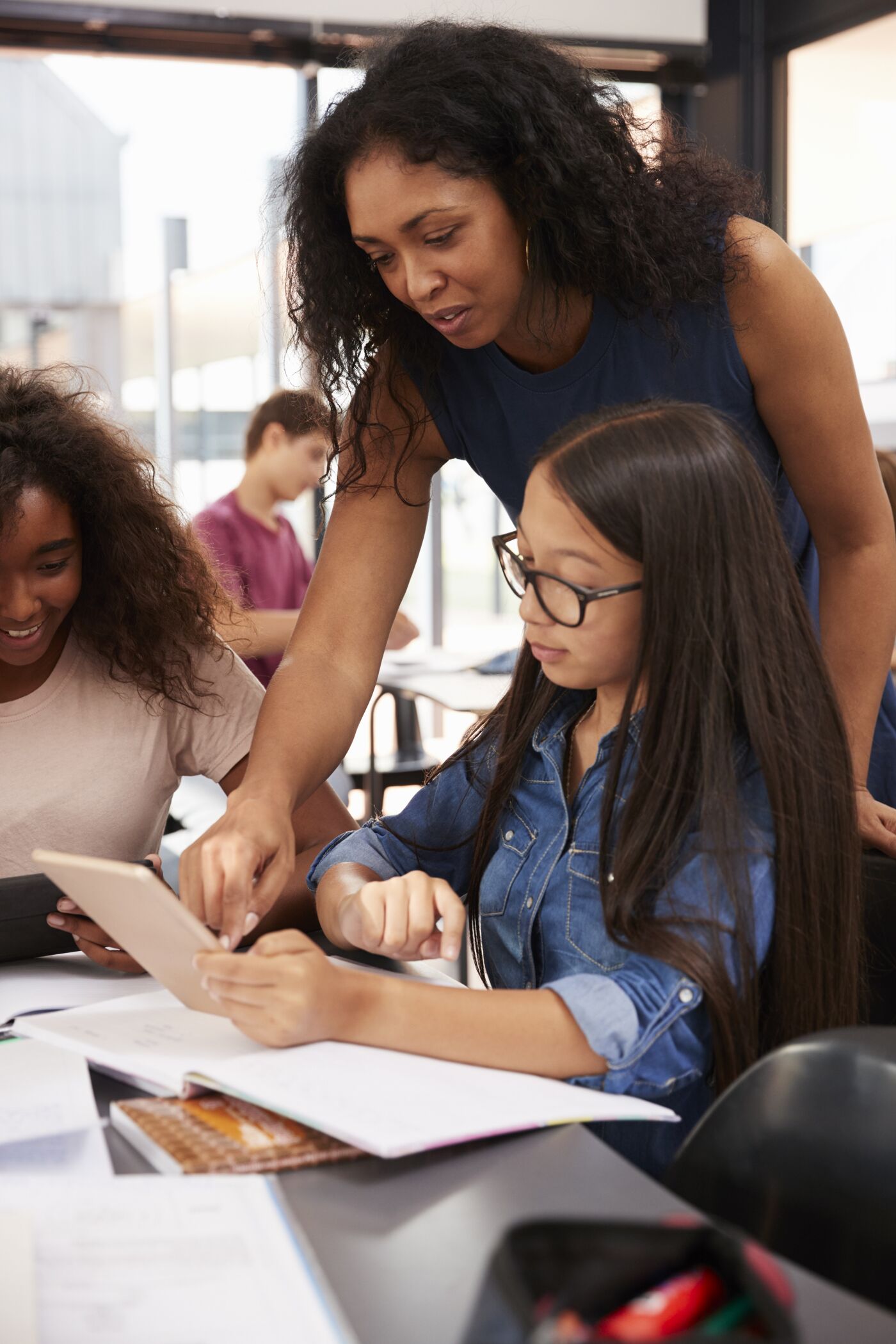 A female teacher leans over to help a young female student using a tablet in a lively classroom setting.