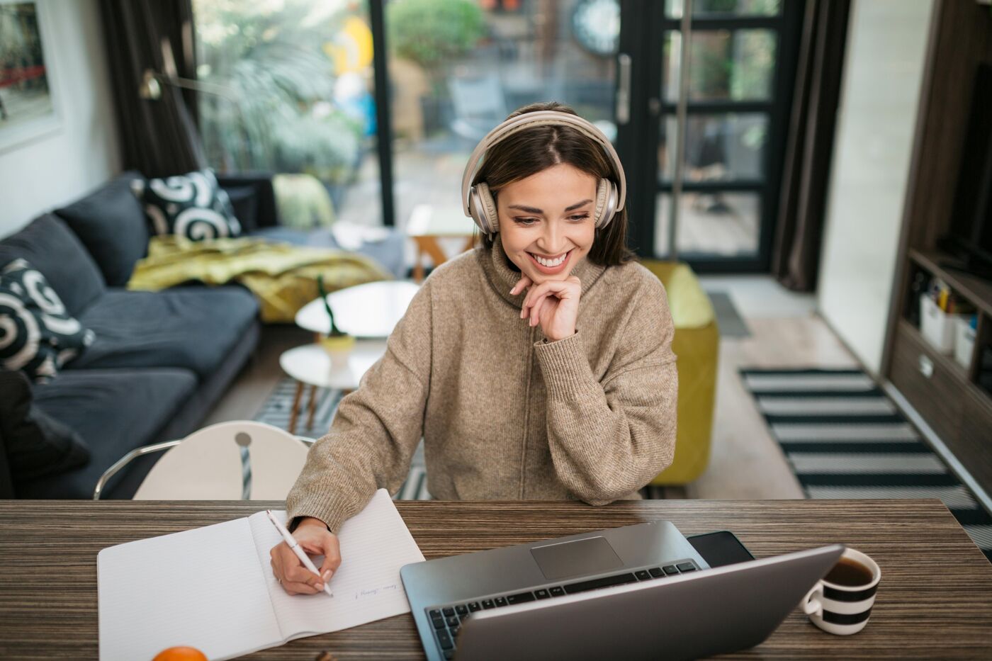 A woman wearing headphones smiles while sitting at a table, working on a laptop and taking notes in a notebook.