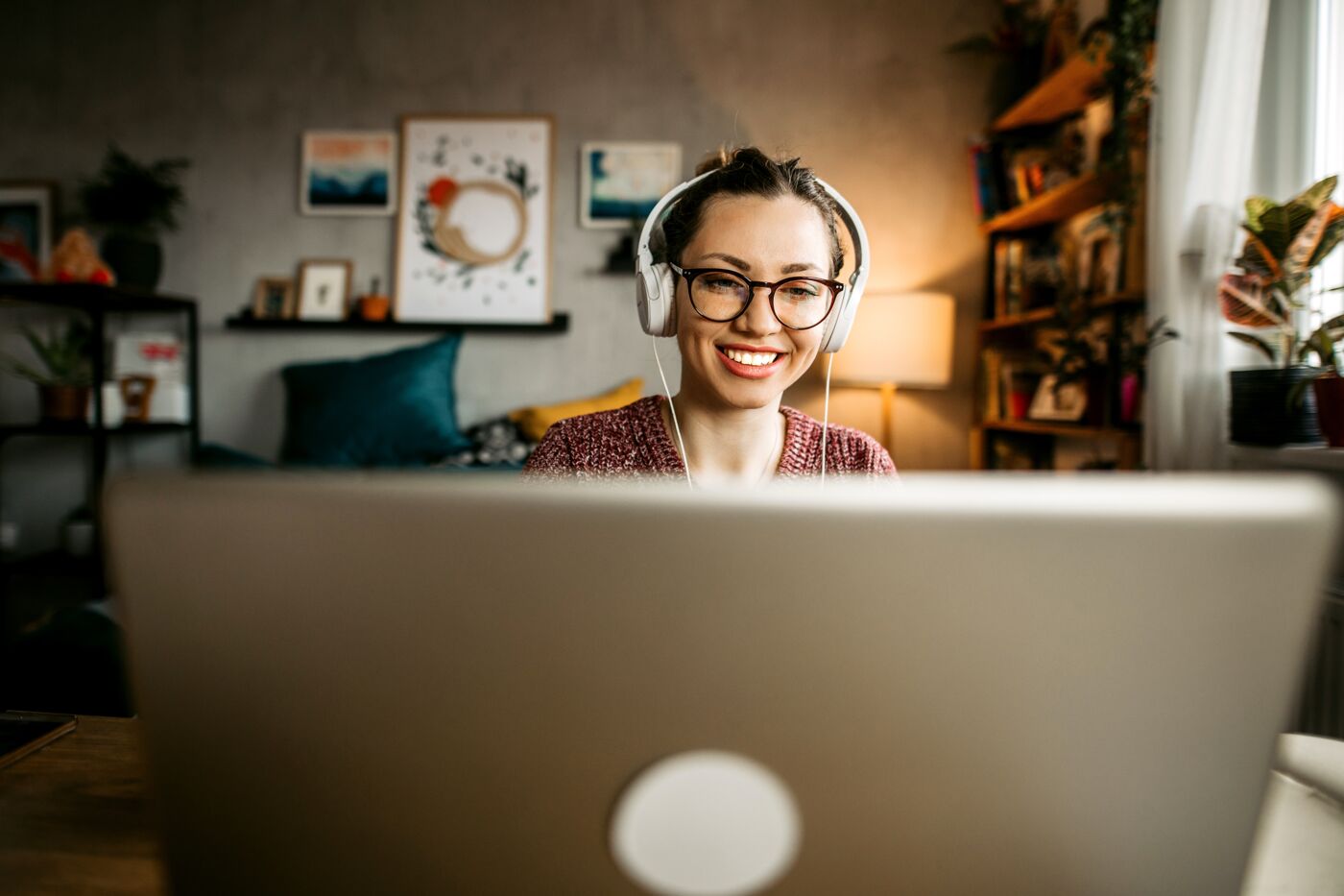 A cheerful woman with headphones working on a laptop in a cozy home office setup, surrounded by warm decor.