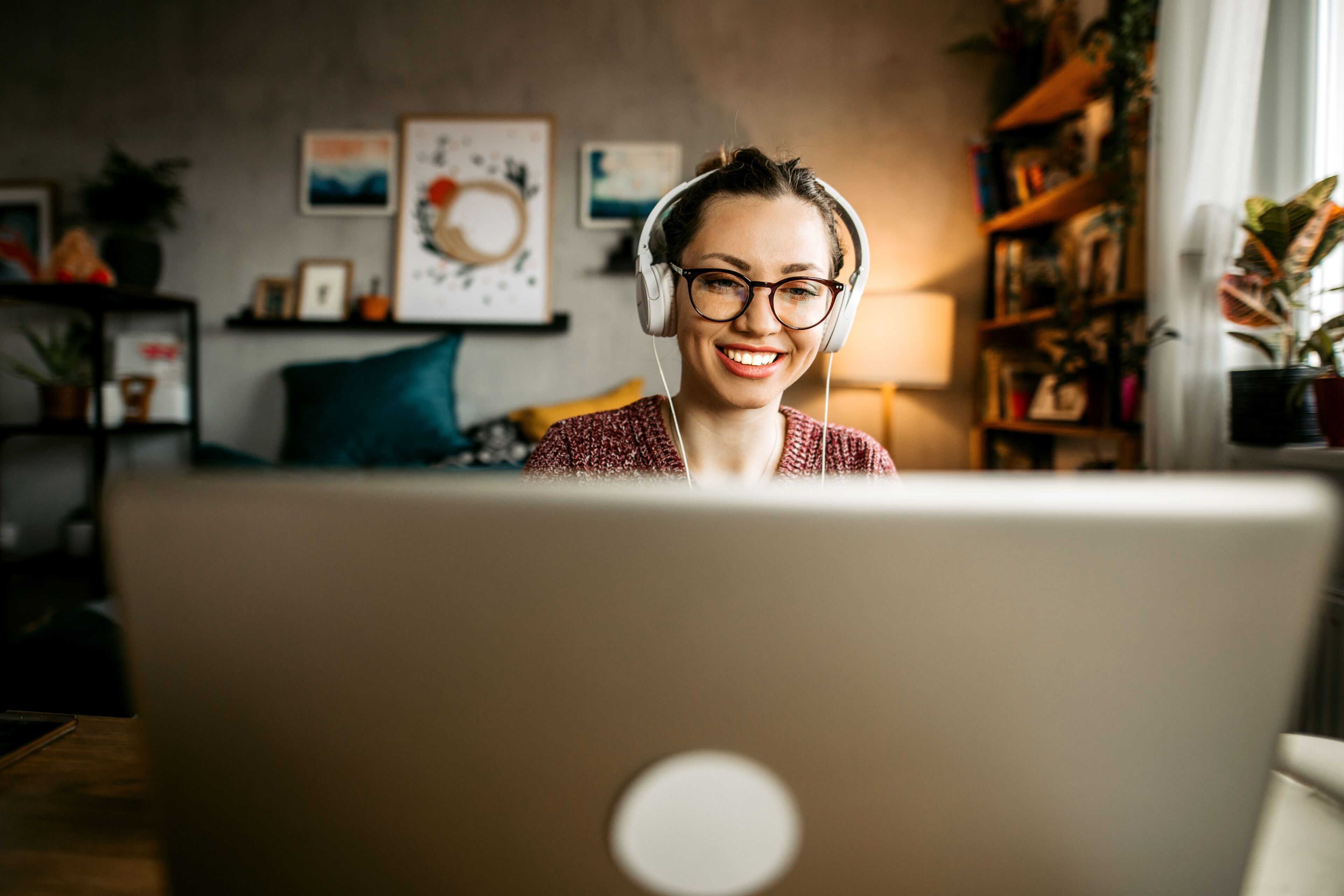 A cheerful woman with headphones working on a laptop in a cozy home office setup, surrounded by warm decor.