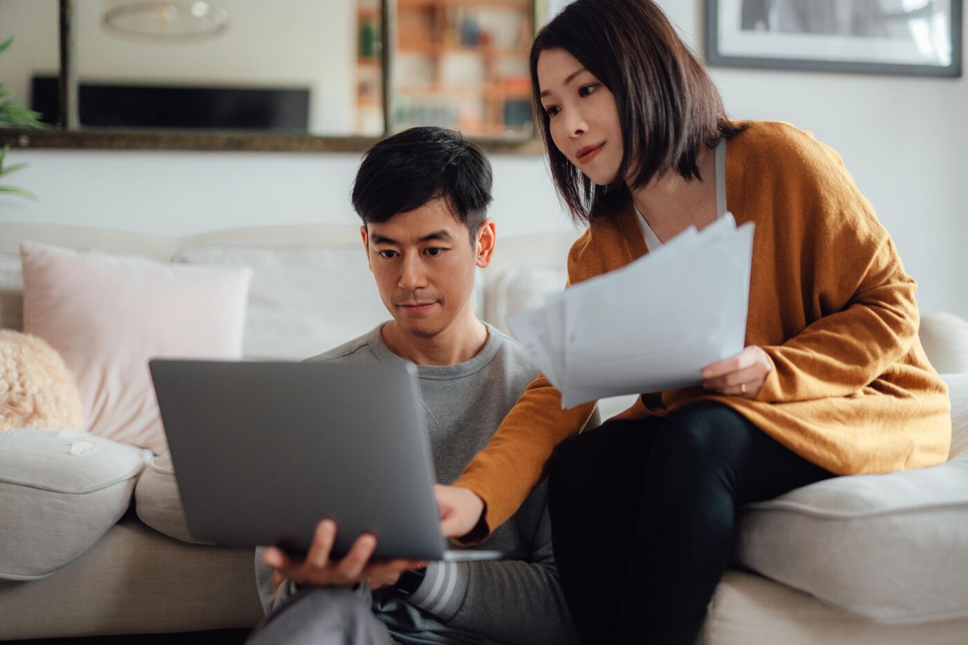 A young couple sits on a couch, looking at a laptop together, with the woman holding some papers.