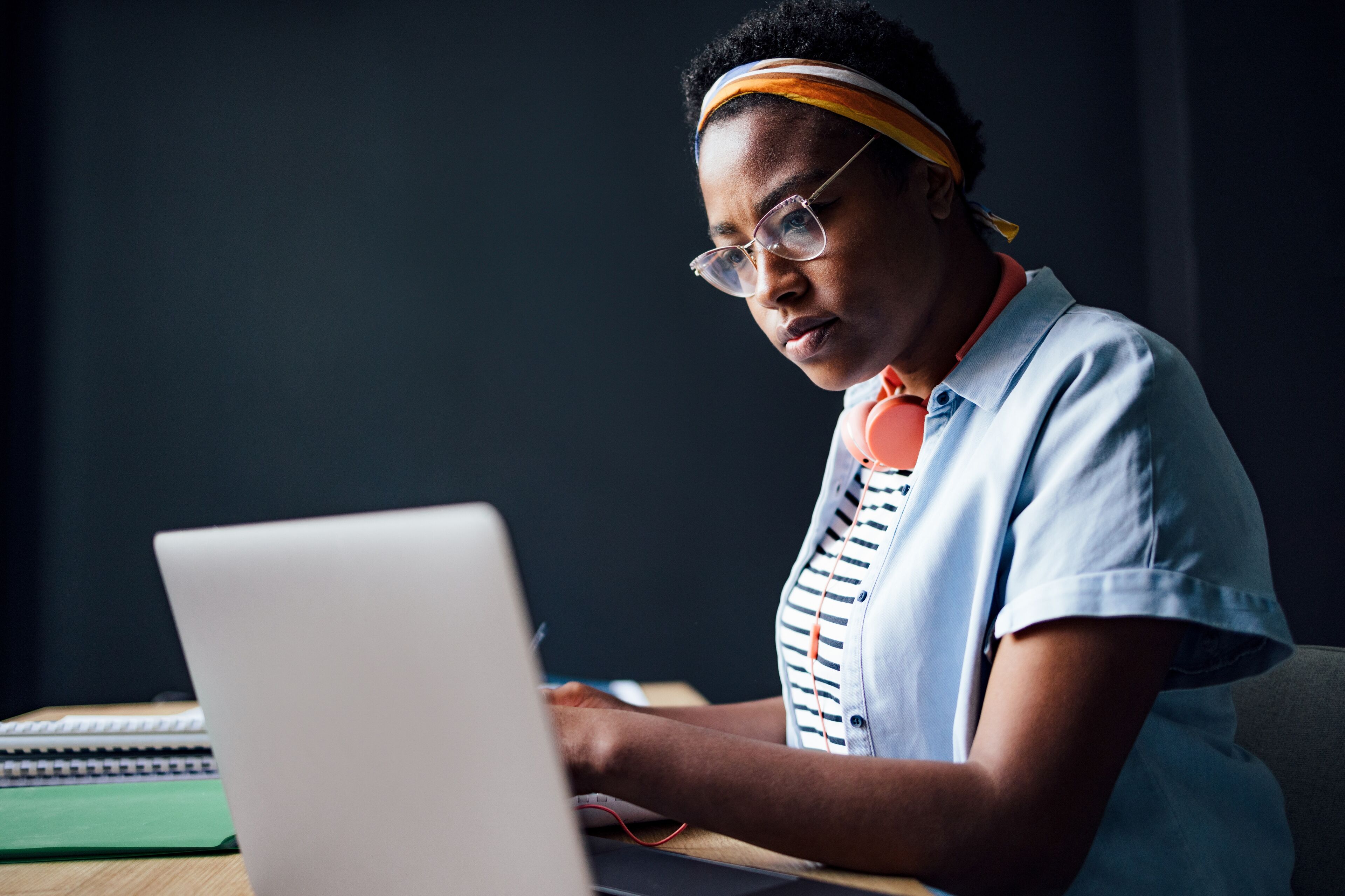 Serious African-American woman studying using a laptop computer while sitting at desk and writing homework in a notebook.