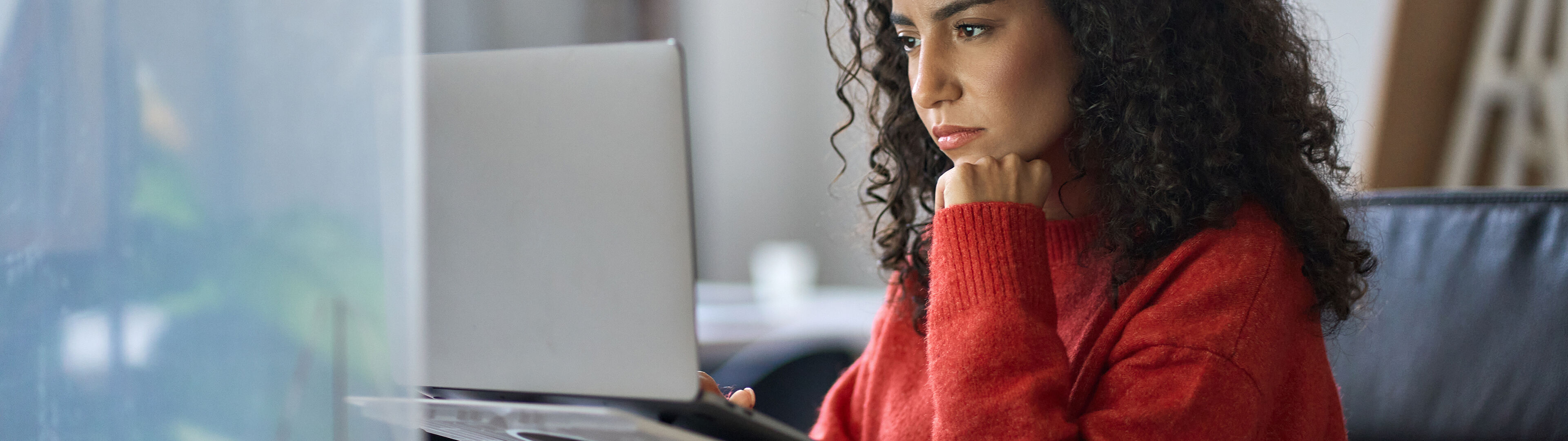 Une femme pensive en pull rouge concentrée sur son travail devant un ordinateur dans un bureau.