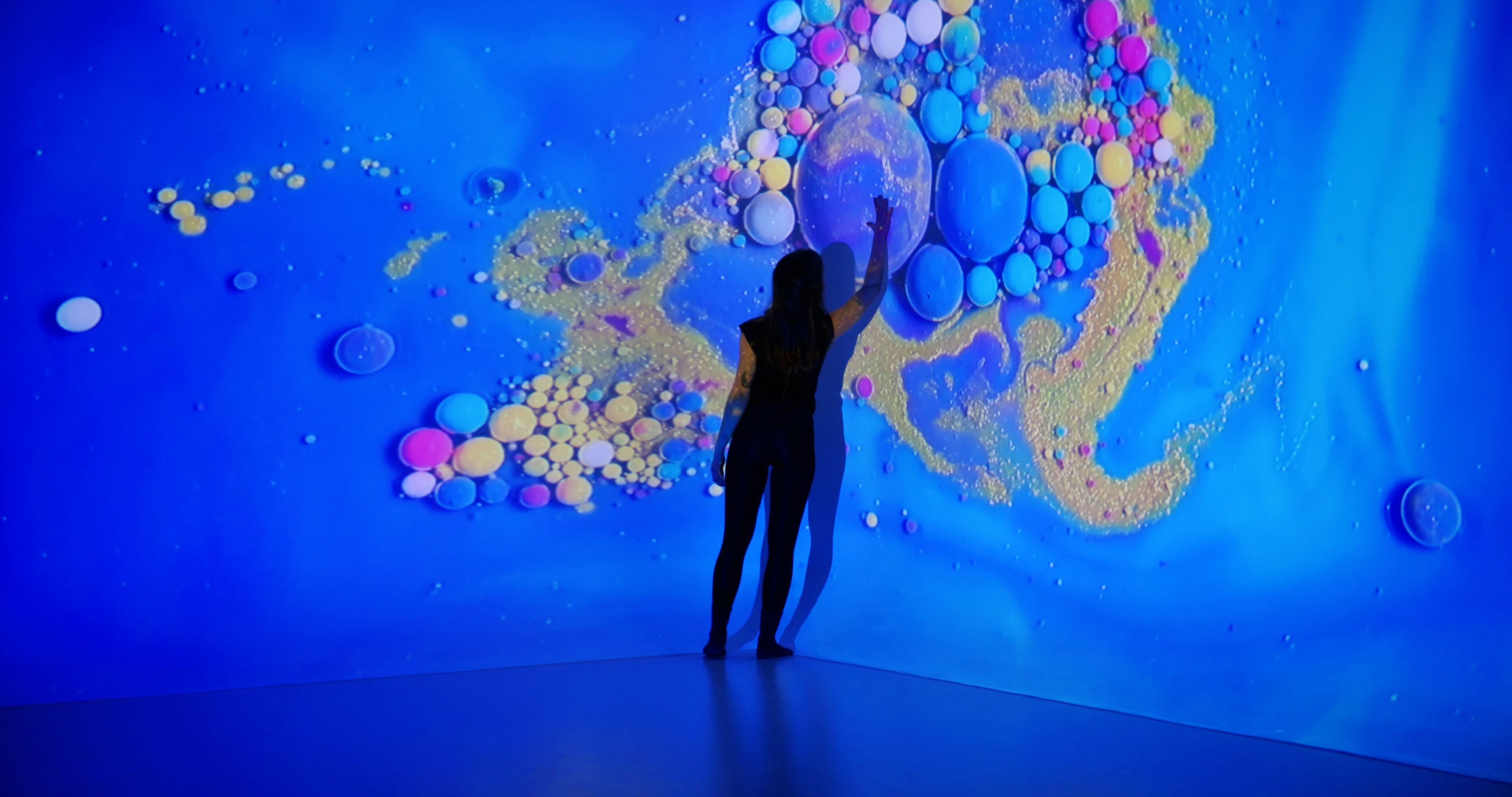 Una persona está silueteada contra una vibrante exposición de arte interactivo con burbujas iridiscentes y dinámicas en un fondo azul intenso.