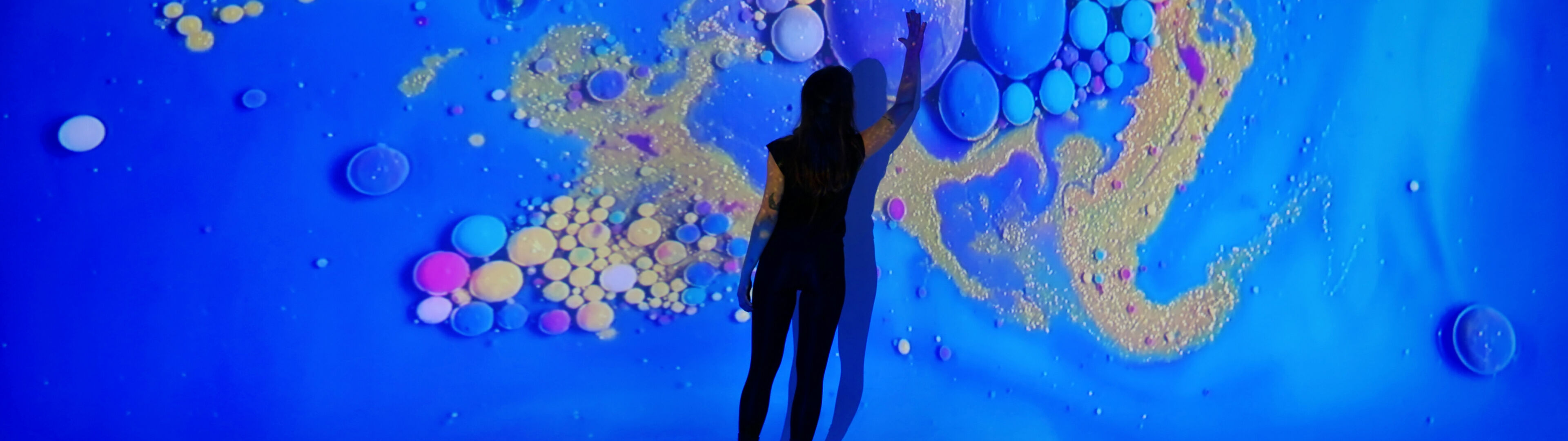 A person is silhouetted against a vibrant, interactive art display featuring dynamic, iridescent bubbles on a deep blue background.