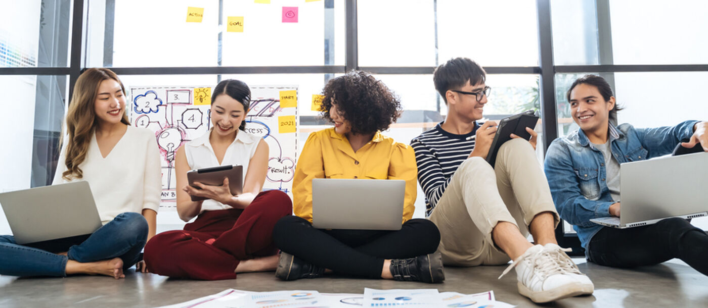 A group of six diverse young professionals are sitting cross-legged on the floor in front of a large window, working on laptops and tablets, seemingly collaborating on a project in a modern office space.