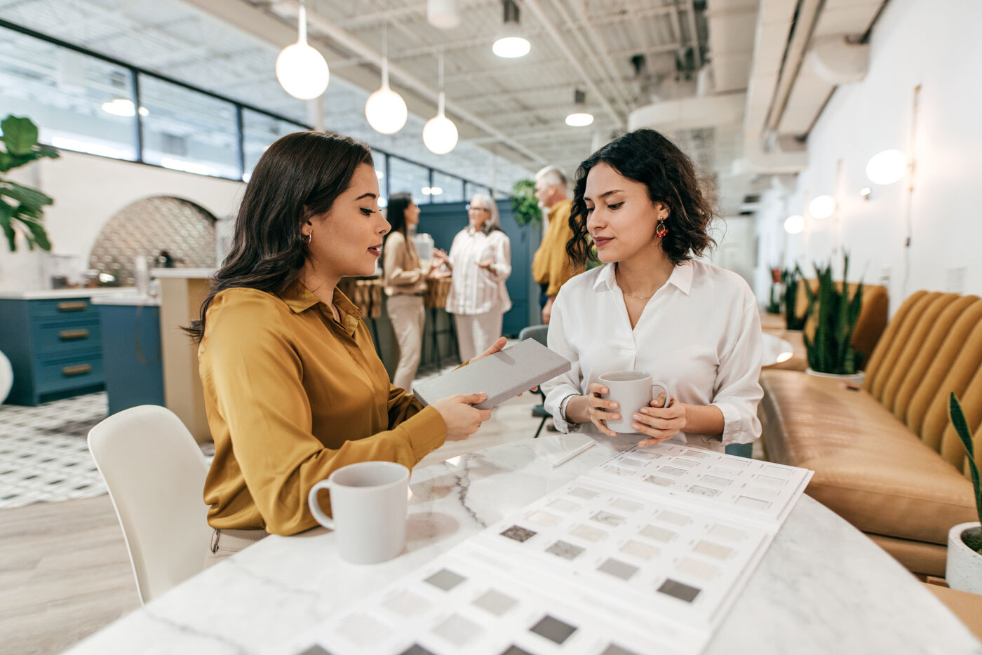 Two women sit at a table in an office, reviewing samples while one holds a notebook and the other holds a mug.