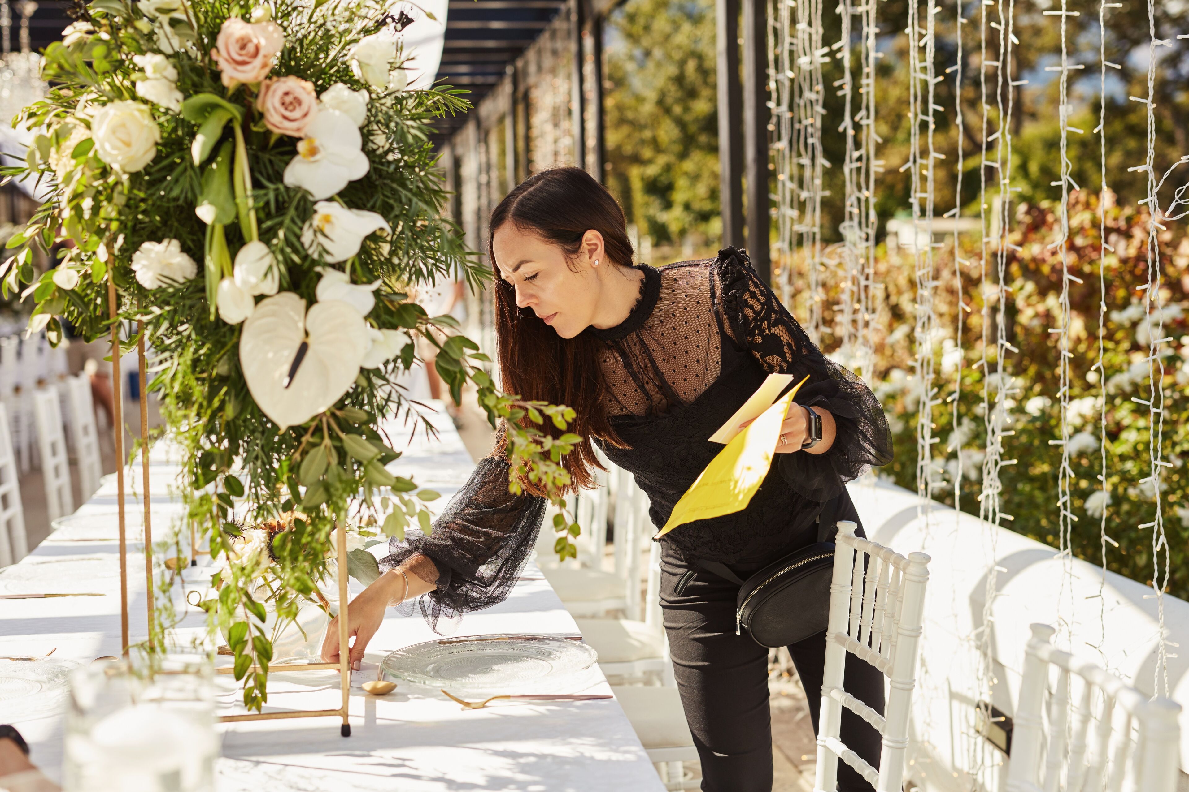 Une fleuriste en blouse noire en dentelle arrange des fleurs blanches et roses sur une table d'événement décorée, concentrée et attentive aux détails.