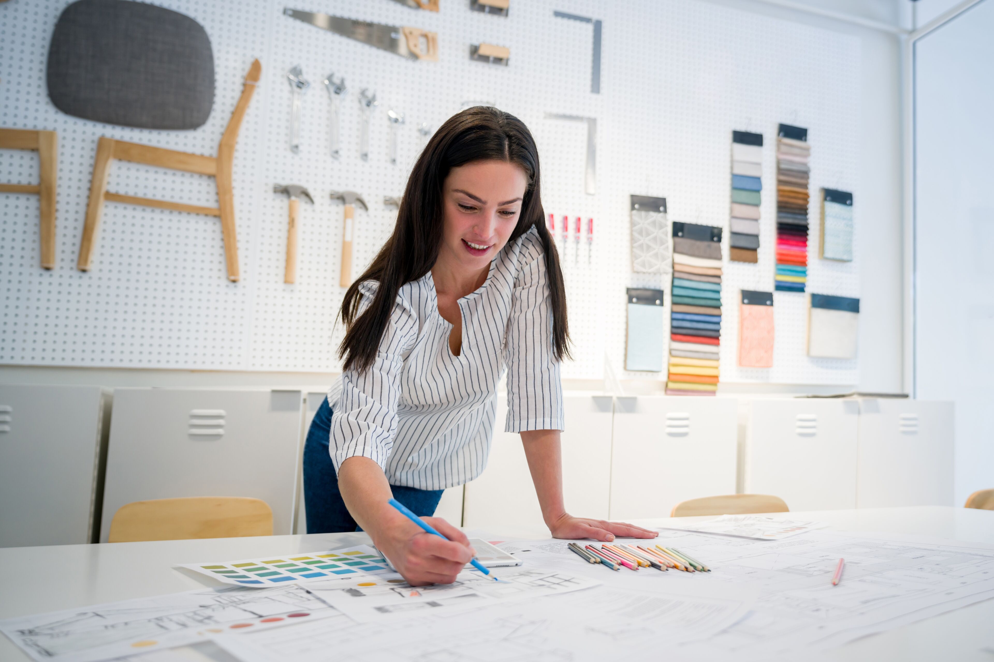 A focused female interior designer sketches on a blueprint in a bright studio, with fabric samples and design tools on the wall behind her.