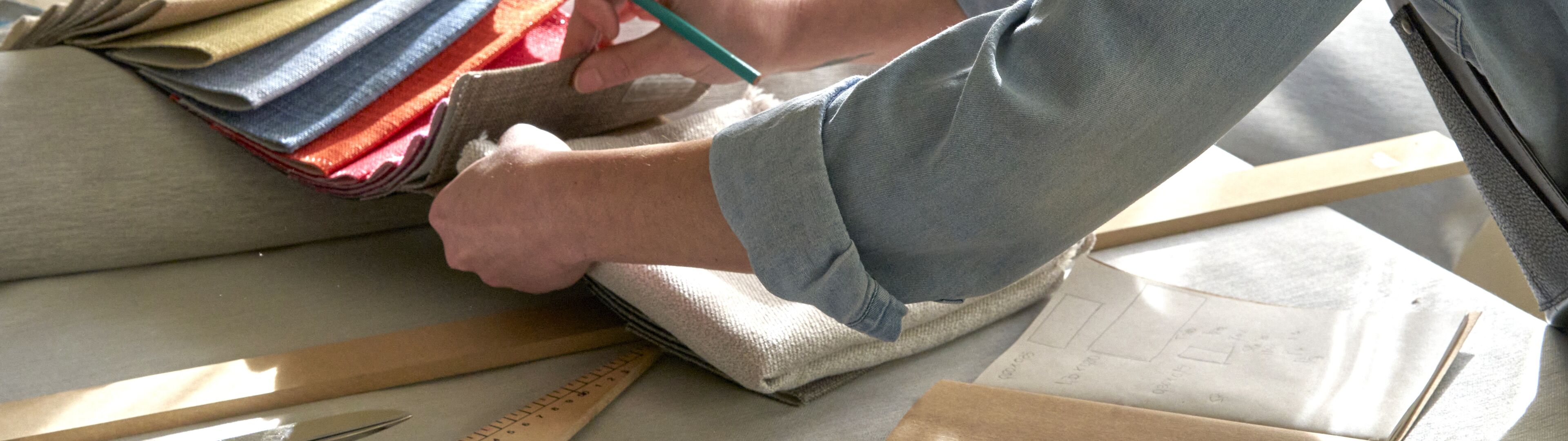 Una persona selecciona meticulosamente muestras de tela para sastrería, con herramientas de medición y patrones sobre una mesa de madera en un espacio de trabajo bien iluminado.
