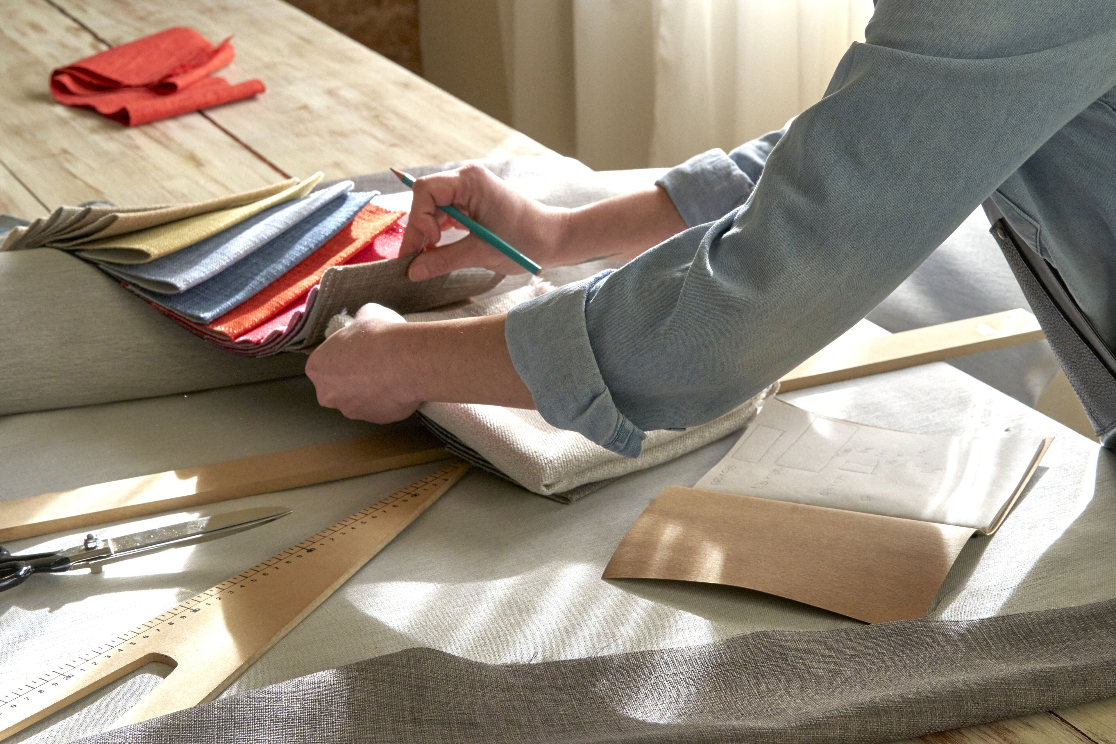 A person meticulously chooses fabric swatches for tailoring, with measuring tools and patterns laid out on a wooden table, in a well-lit workspace.