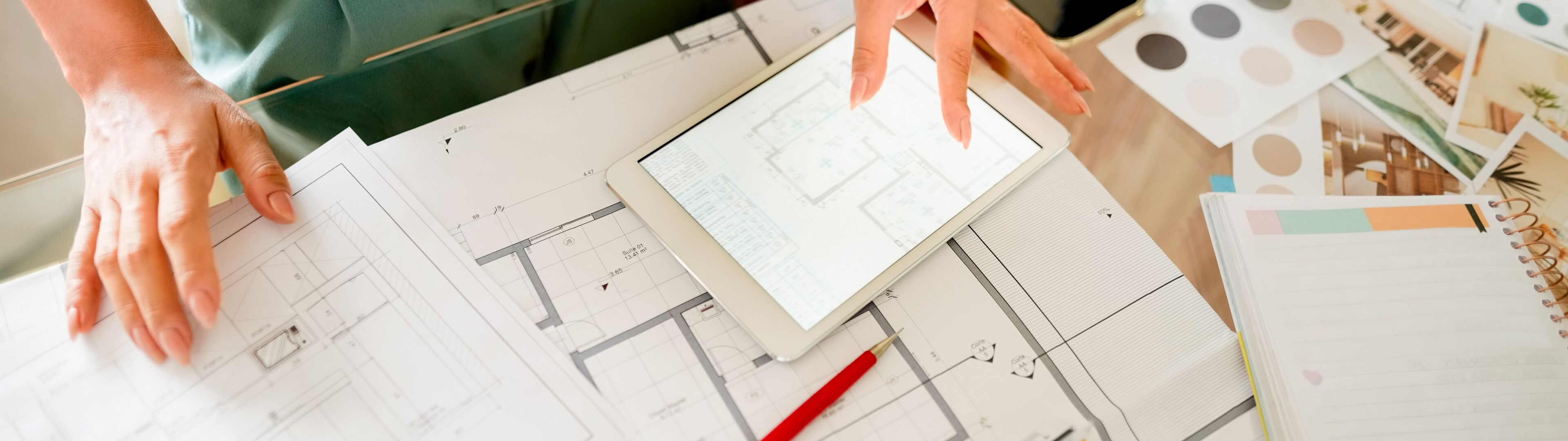 A professional reviewing architectural plans on a tablet over a desk cluttered with design drawings and color palettes.