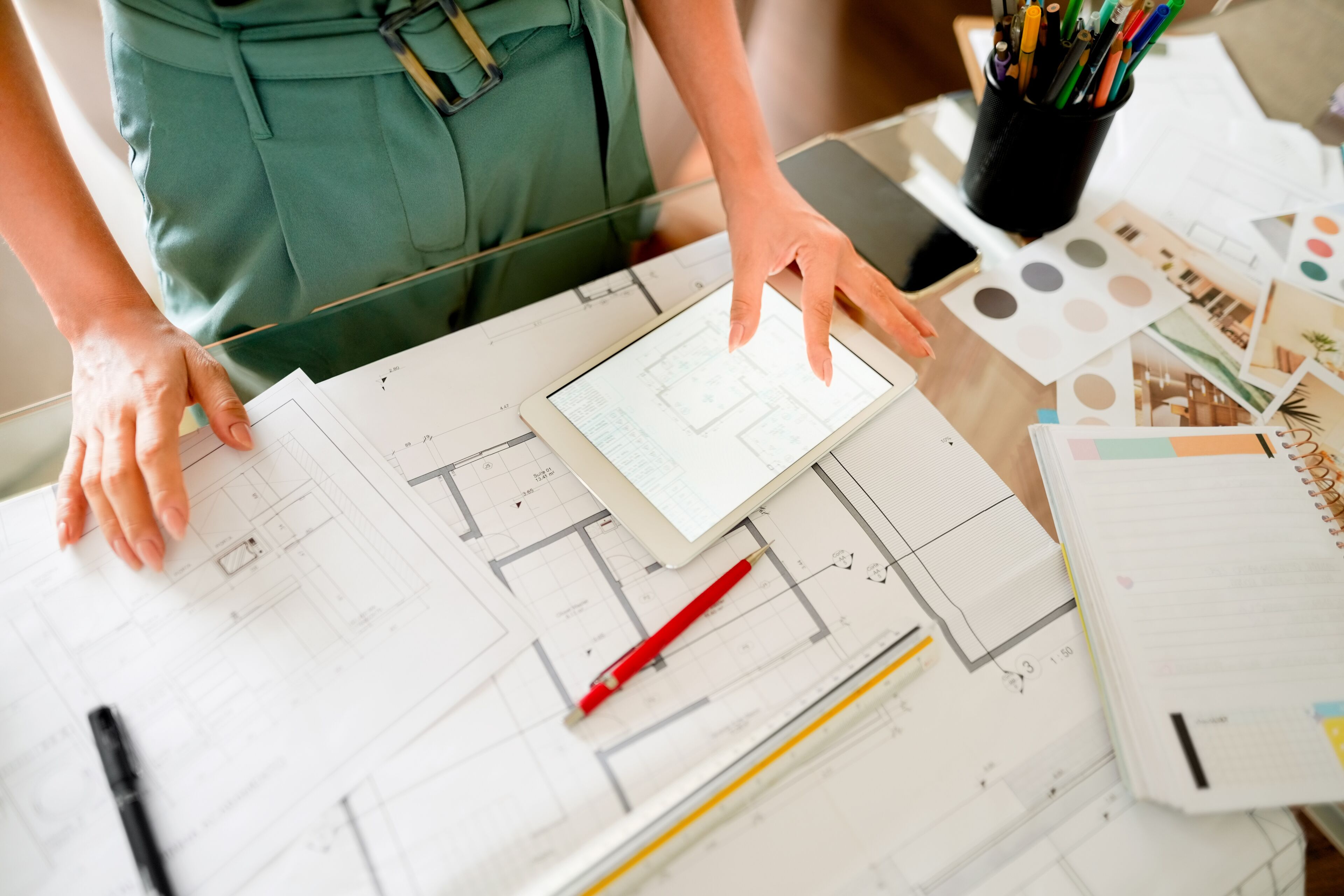 Un/a professional revisa plans arquitectònics en una tauleta sobre un escriptori ple de dibuixos de disseny i paletes de colors.