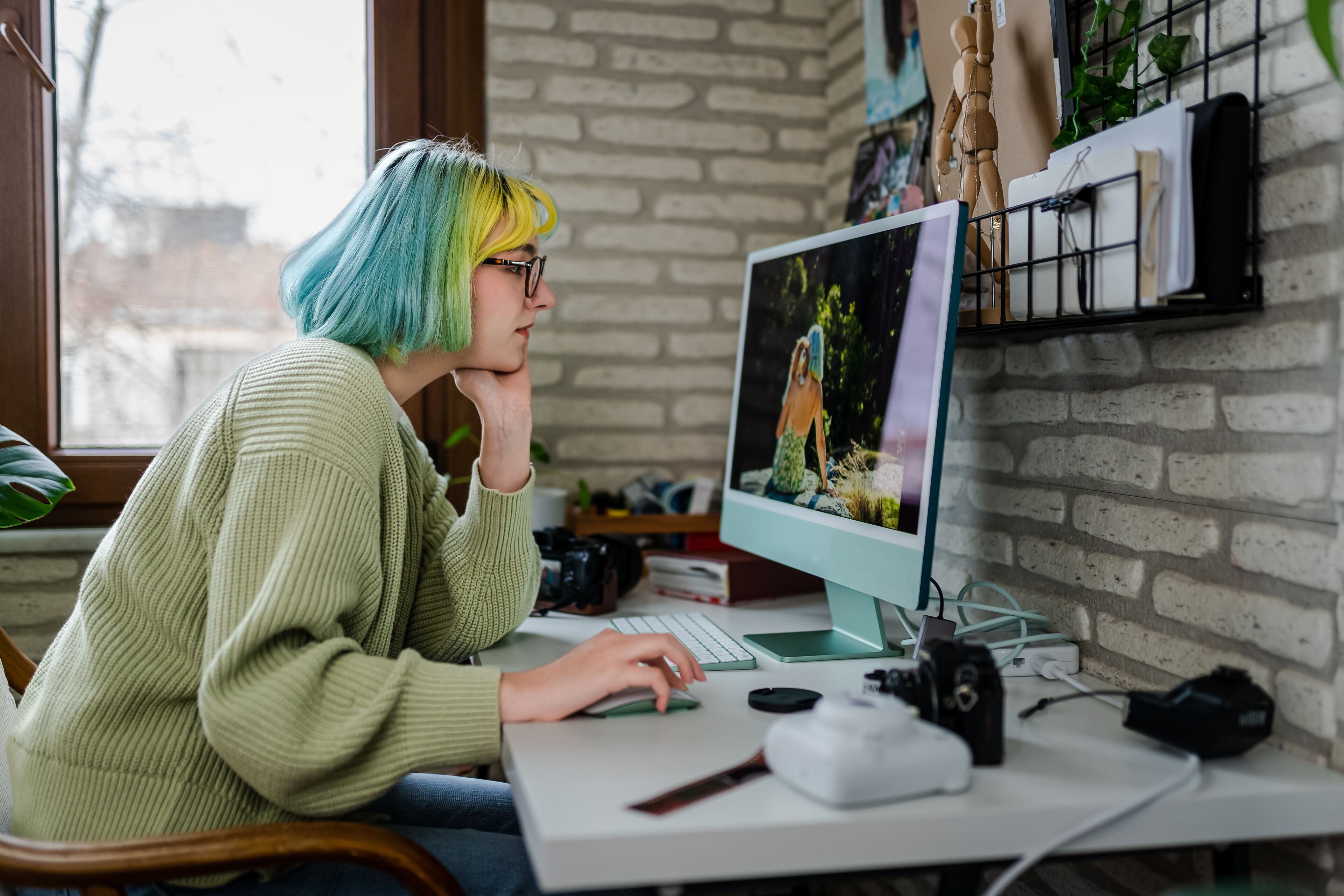 Una persona amb cabells de colors variats està immersa en la seva feina en un ordinador amb equip fotogràfic al voltant.