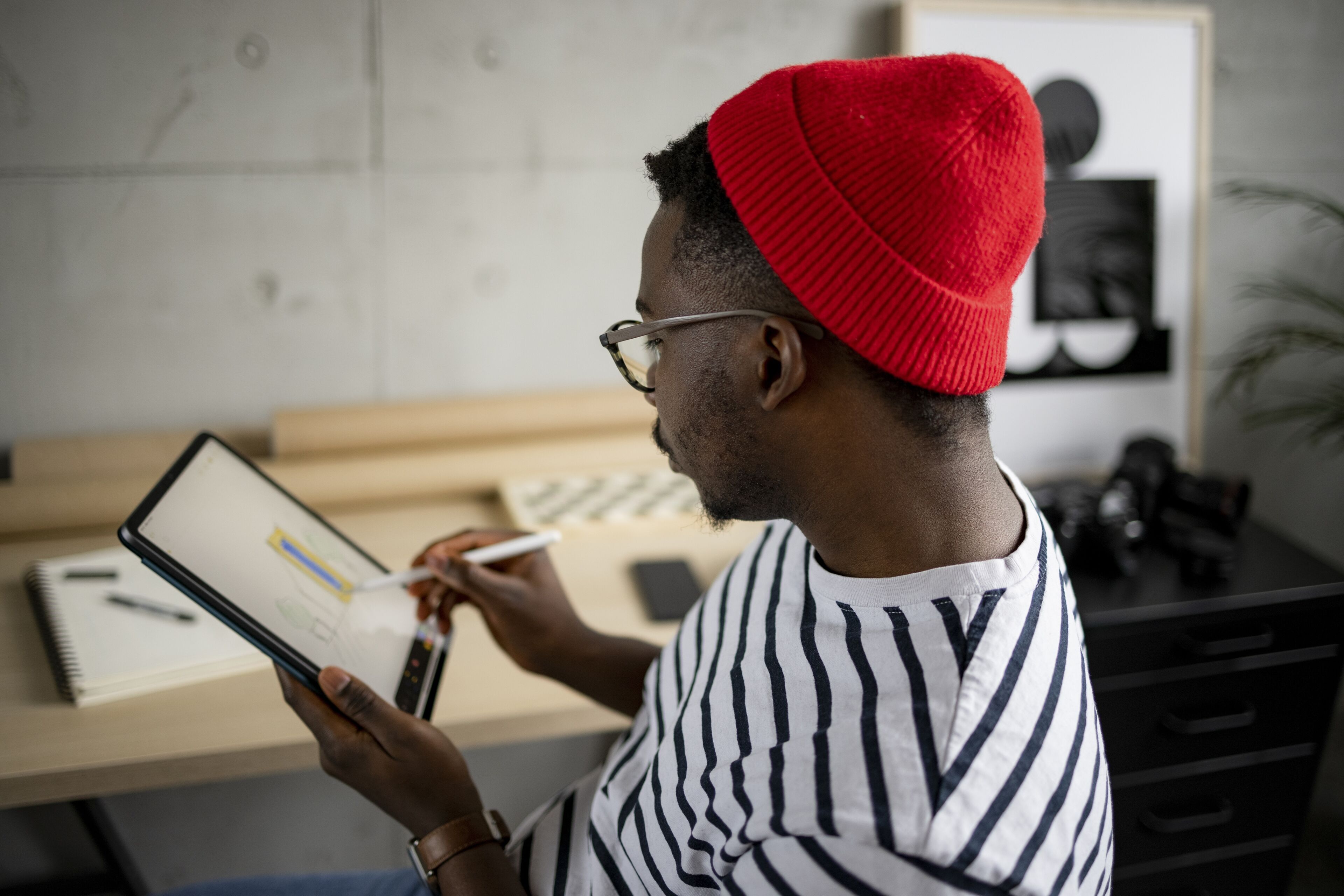 Un homme concentré avec un bonnet rouge et une chemise rayée utilise un stylet sur une tablette numérique dans un espace de travail moderne.

