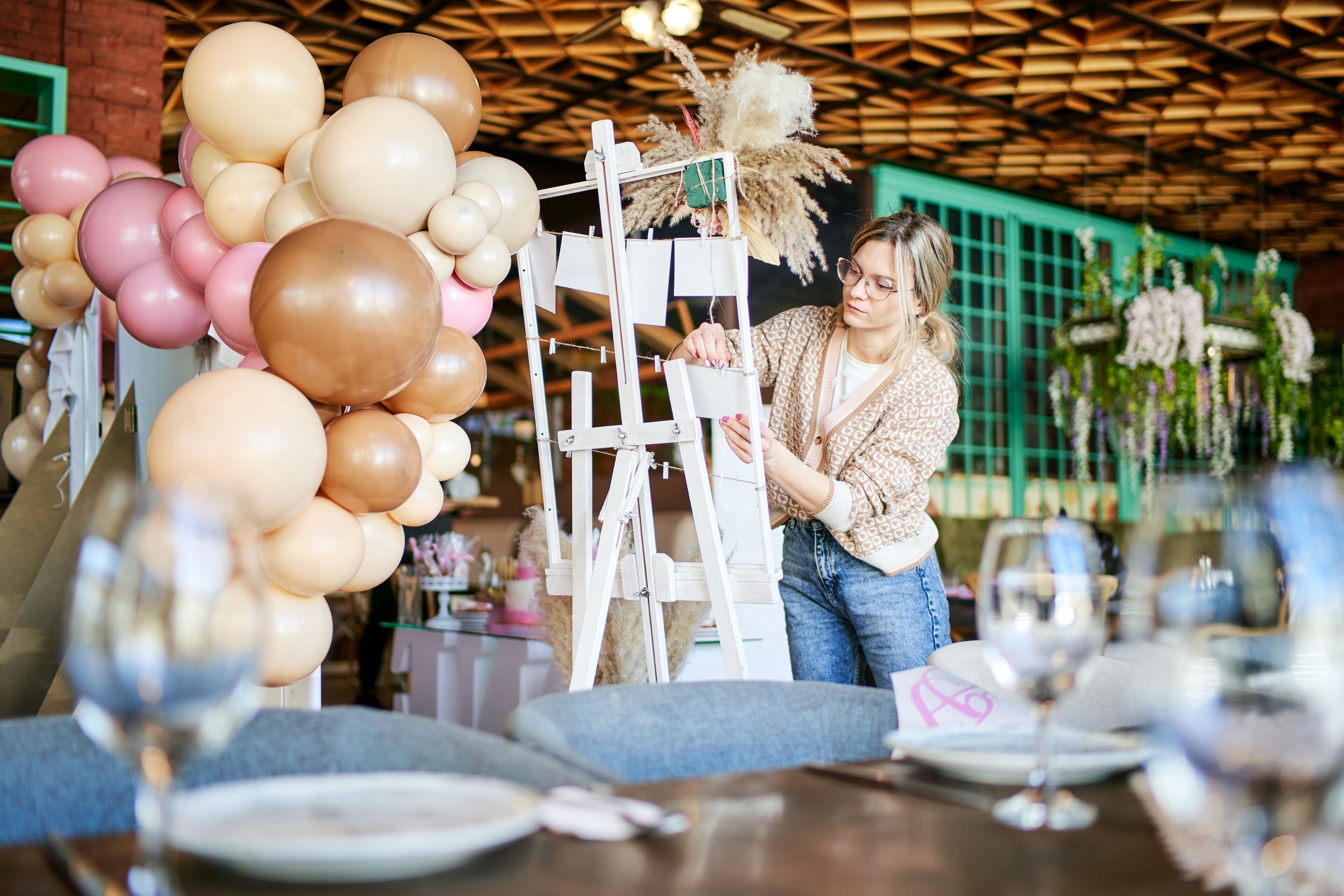 Une organisatrice d'événements concentrée ajuste les décorations sur un chevalet blanc dans un cadre festif avec des ballons et des tables élégamment dressées.
