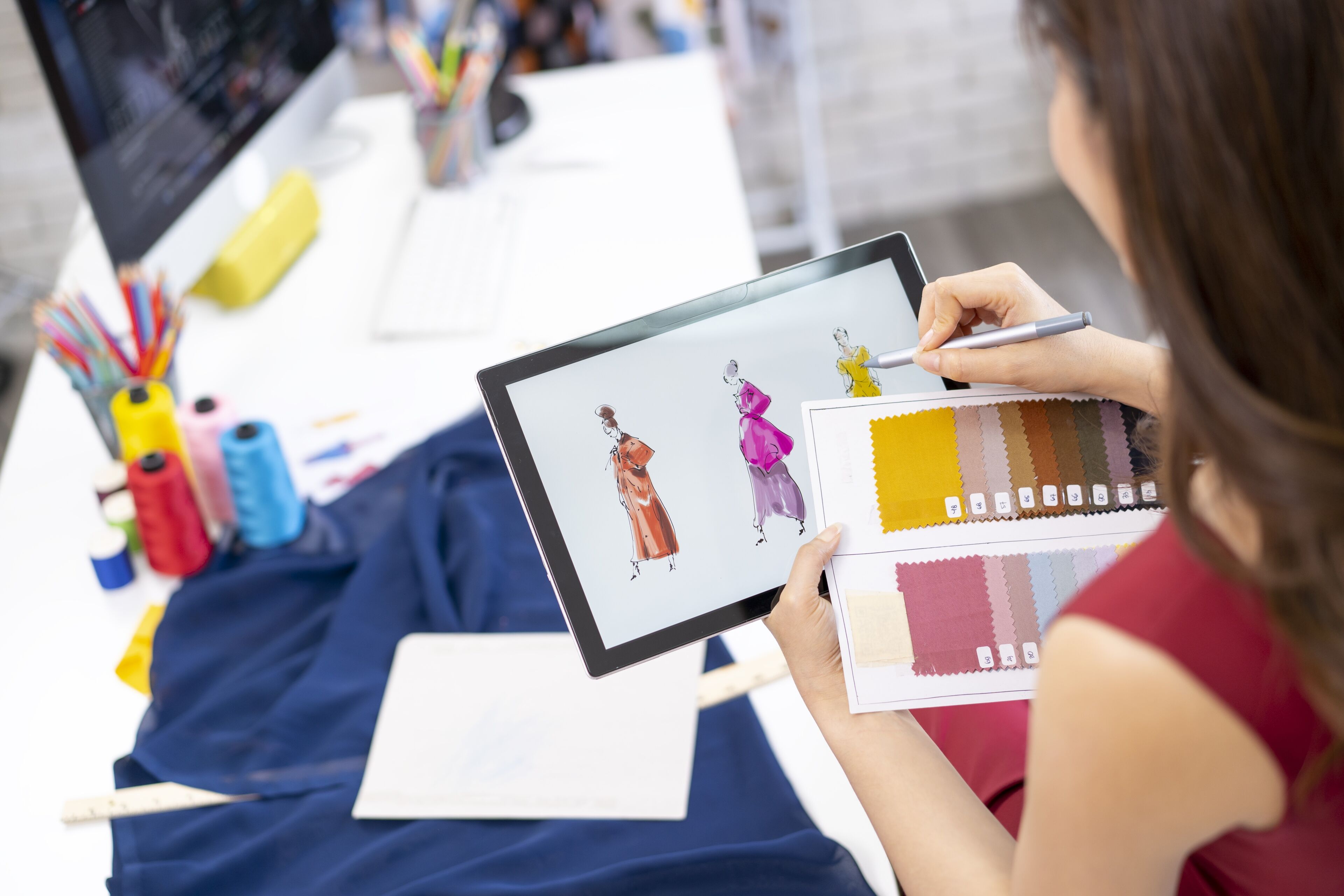 ne créatrice observe des croquis numériques de vêtements sur une tablette, en se référant à une palette d'échantillons de tissus, dans un espace de création.