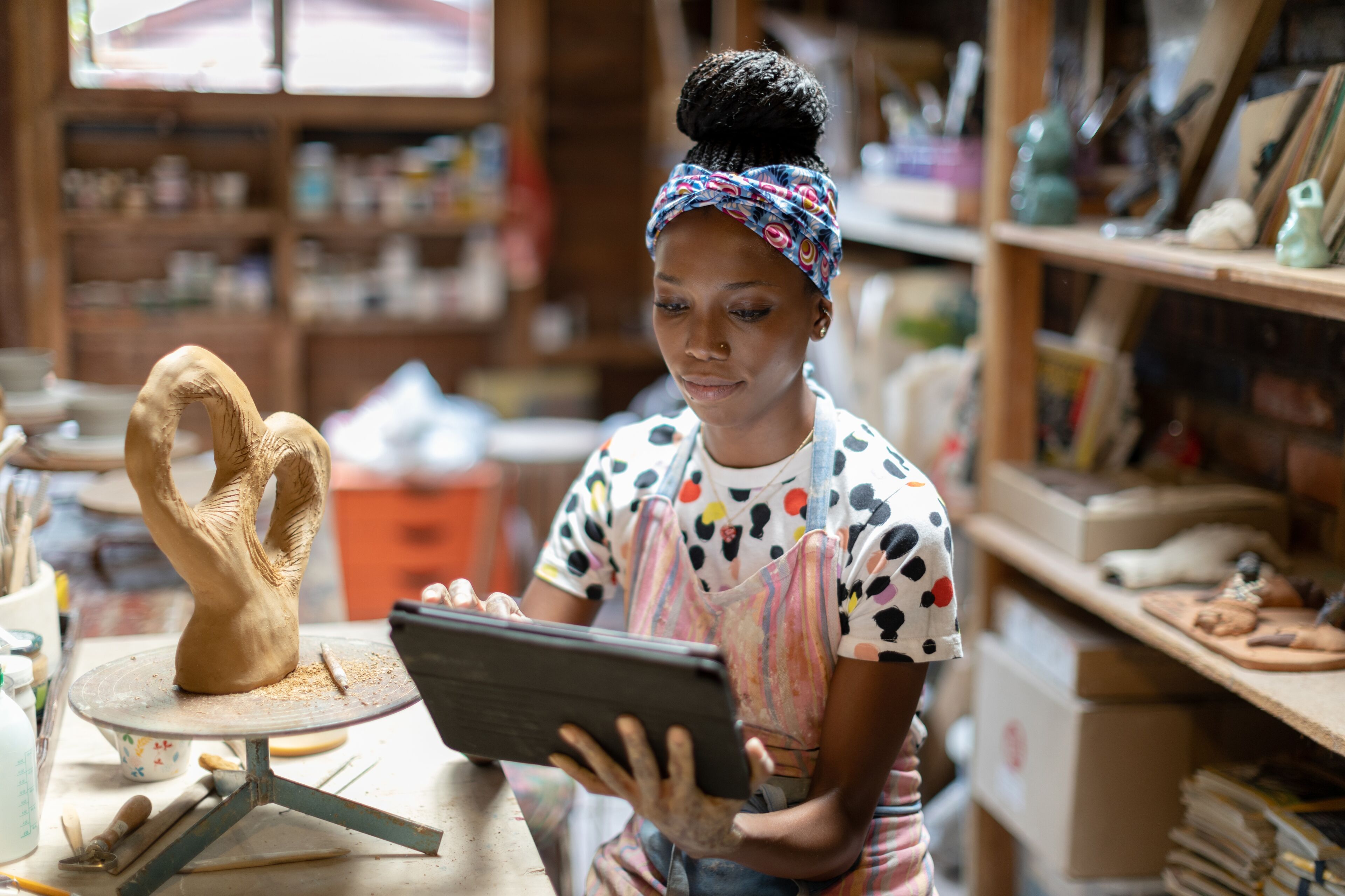 A focused female potter reviews a digital tablet, with her clay sculpture in the foreground, in a cozy, tool-filled workshop.