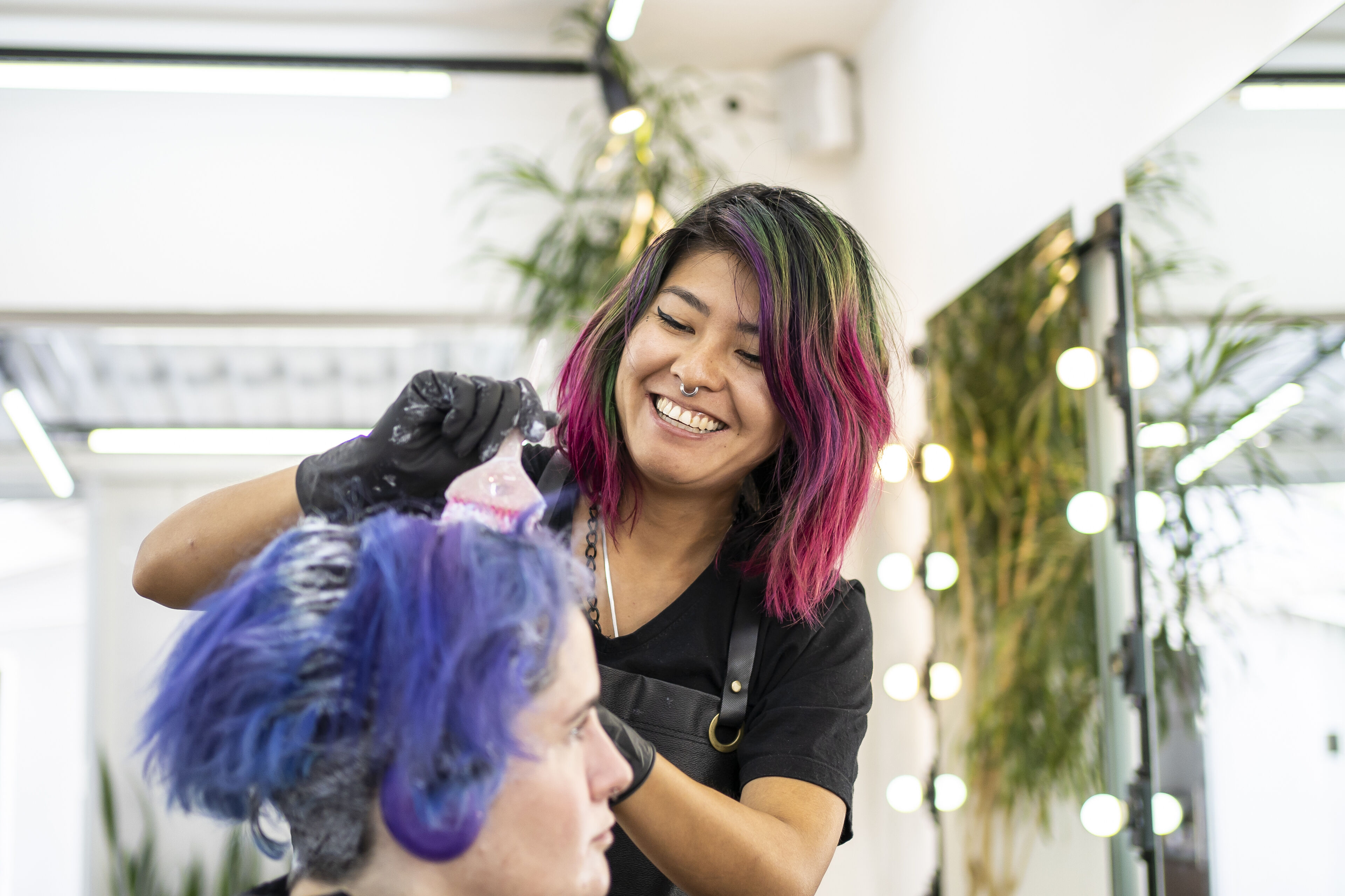 ImageUna colorista de cabello sonriente con cabello rosa vibrante aplica tinte al cabello azul de un cliente en una peluquería iluminada.