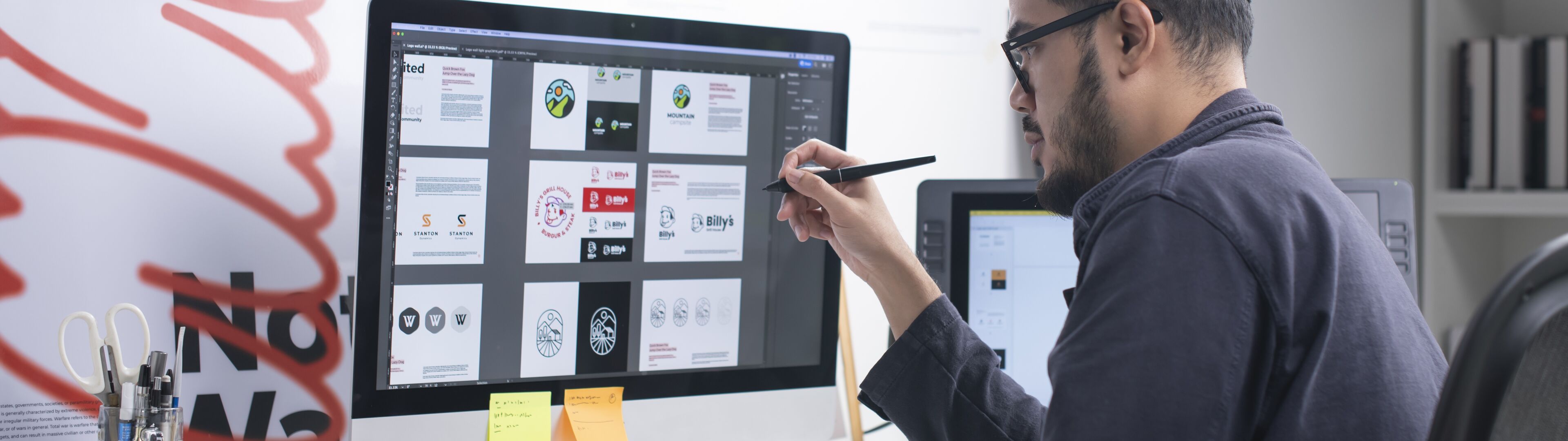 A male graphic designer wearing a beanie reviews designs on a large monitor, stylus in hand, in a well-organized workspace.
