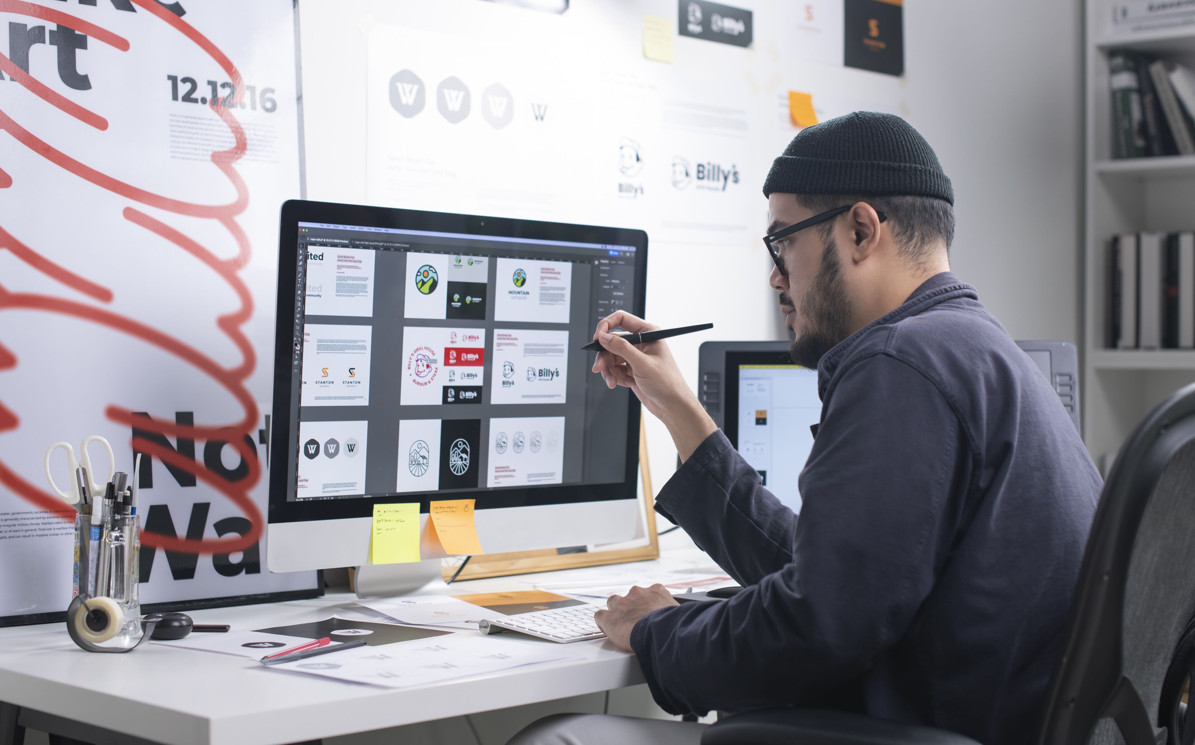 Un graphiste masculin portant un bonnet examine des conceptions sur un grand écran, stylet en main, dans un espace de travail bien organisé.