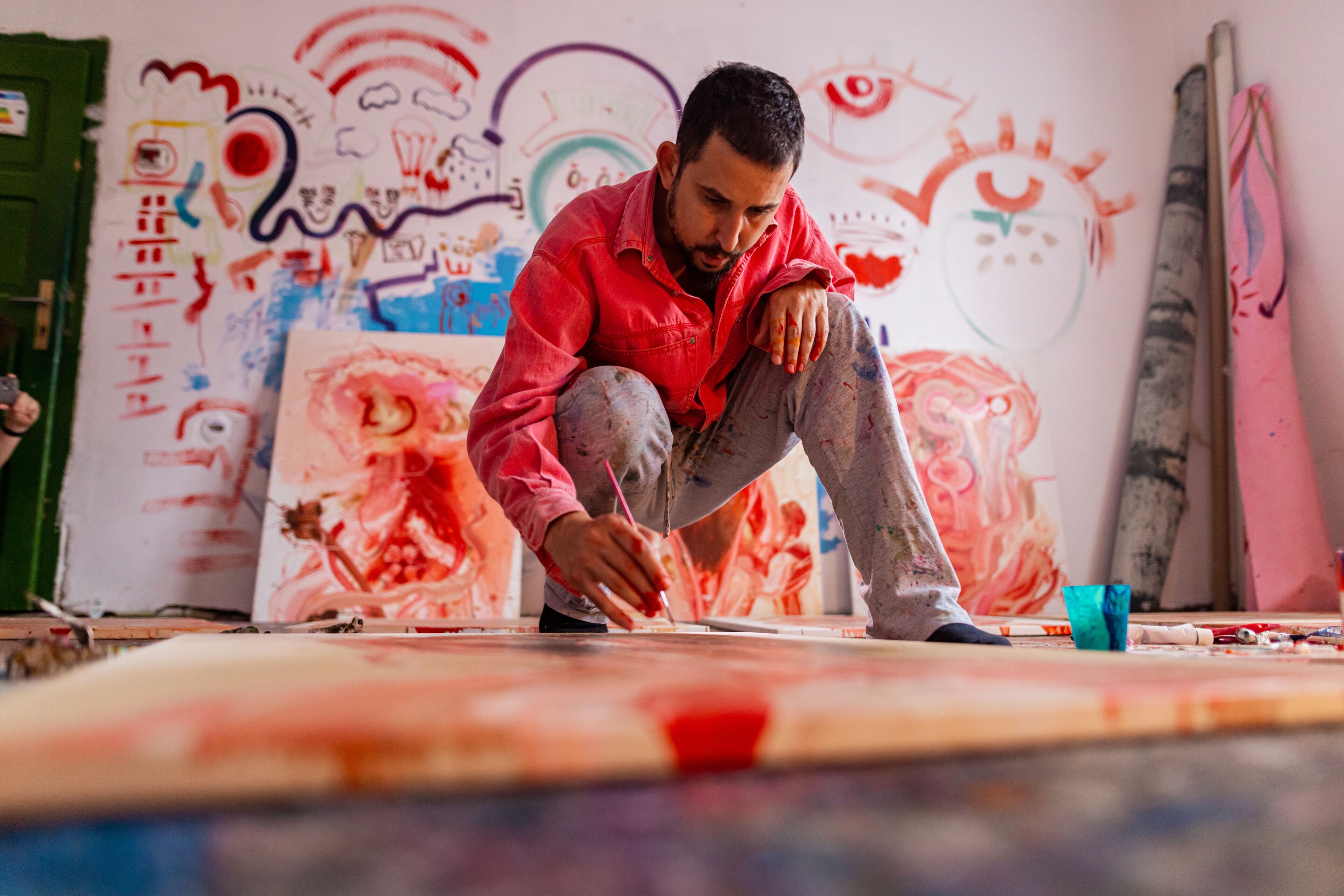 An artist in a red shirt is intently painting on canvas, surrounded by colorful abstract art and a wall filled with whimsical doodles.