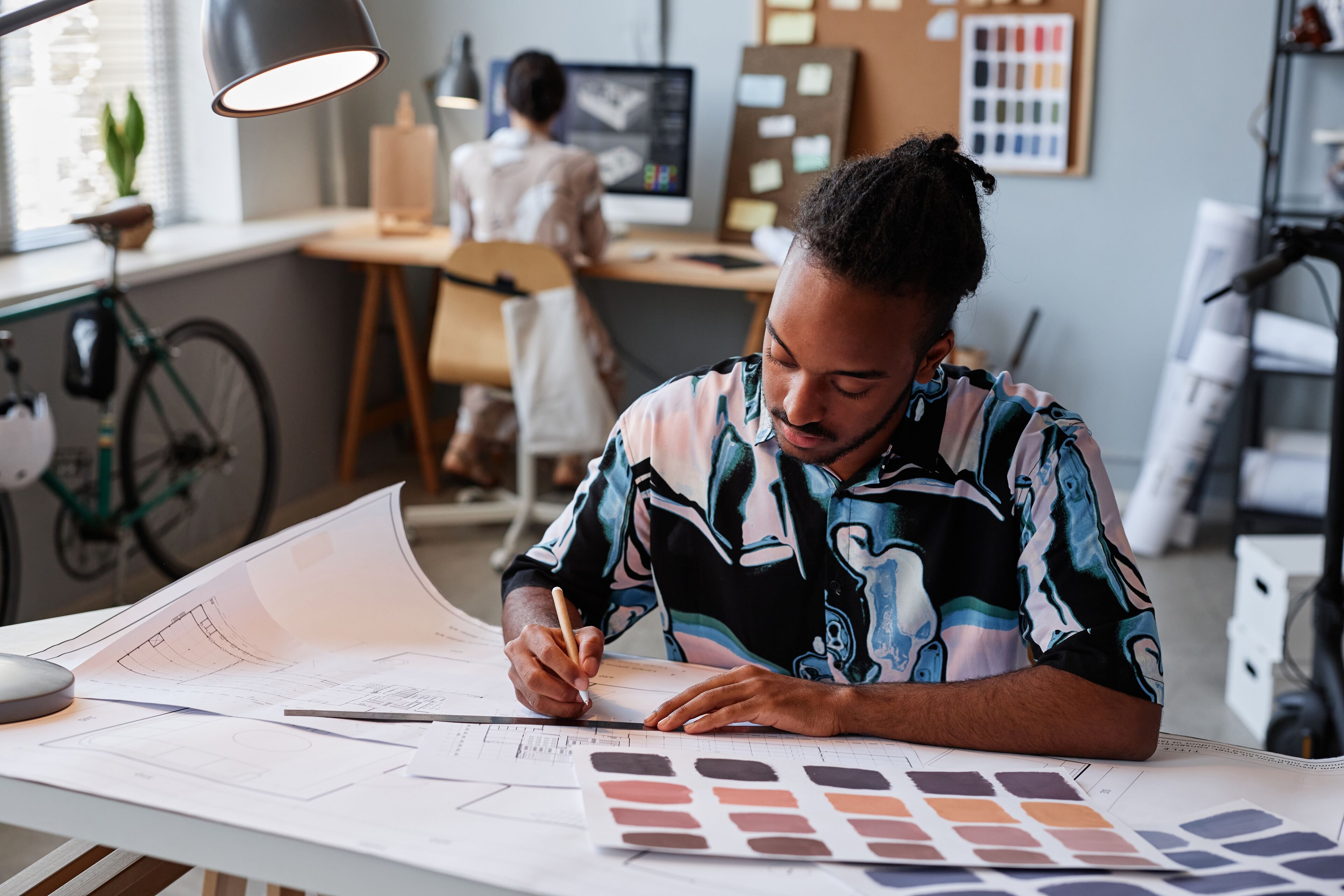 A focused male interior designer drafts plans with color swatches on his desk, with a colleague in the background.