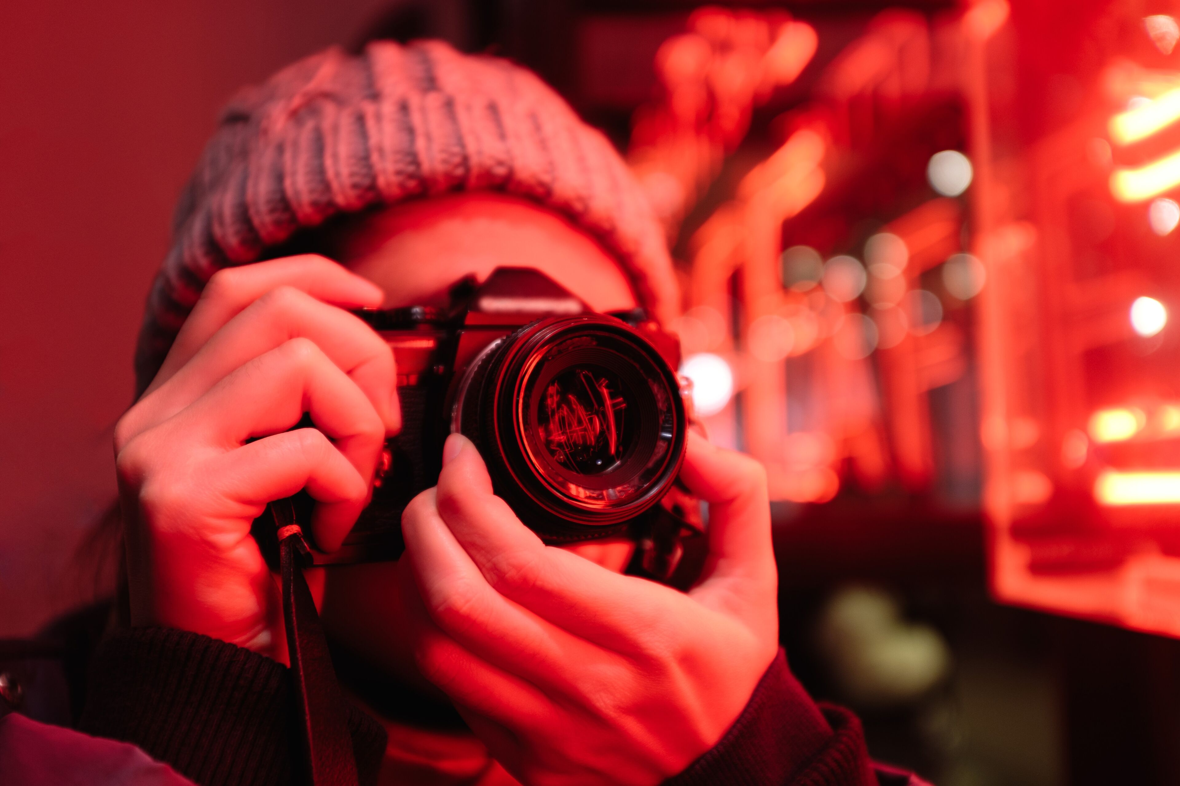 A person wearing a knit hat is taking a photo with a DSLR camera, illuminated by vibrant red neon lighting.