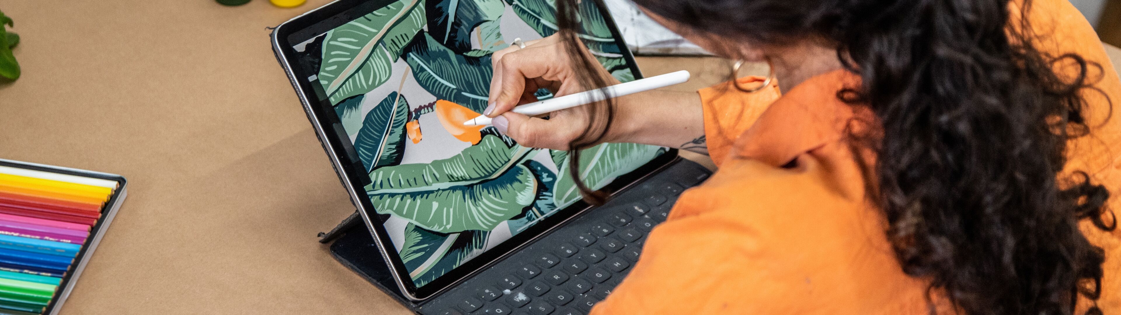 Un artista con camisa naranja usa un lápiz óptico en una tableta para pintar digitalmente un diseño de hojas tropicales.